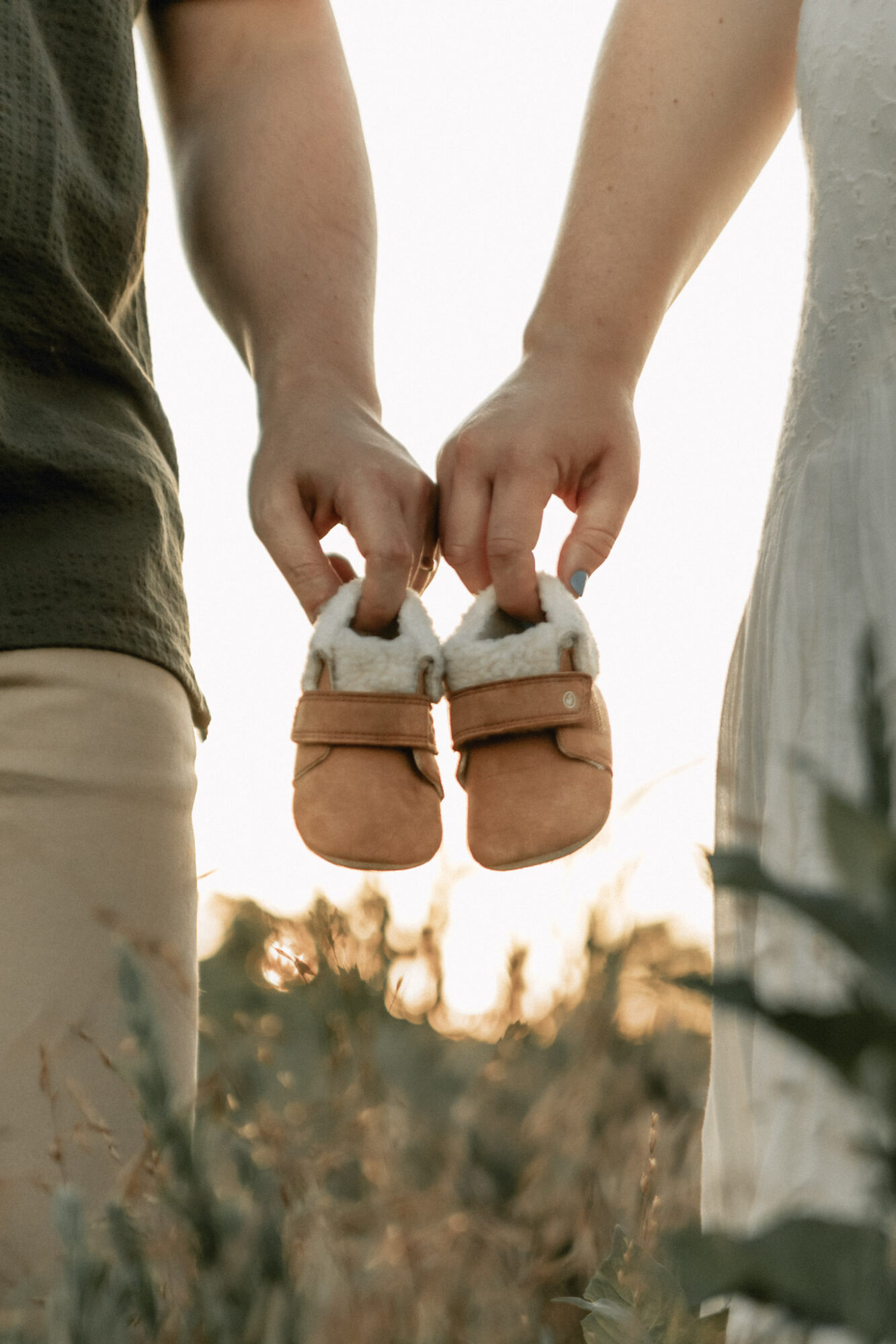 Two people hold small baby shoes between their hands, with a blurred outdoor background and sunlight.