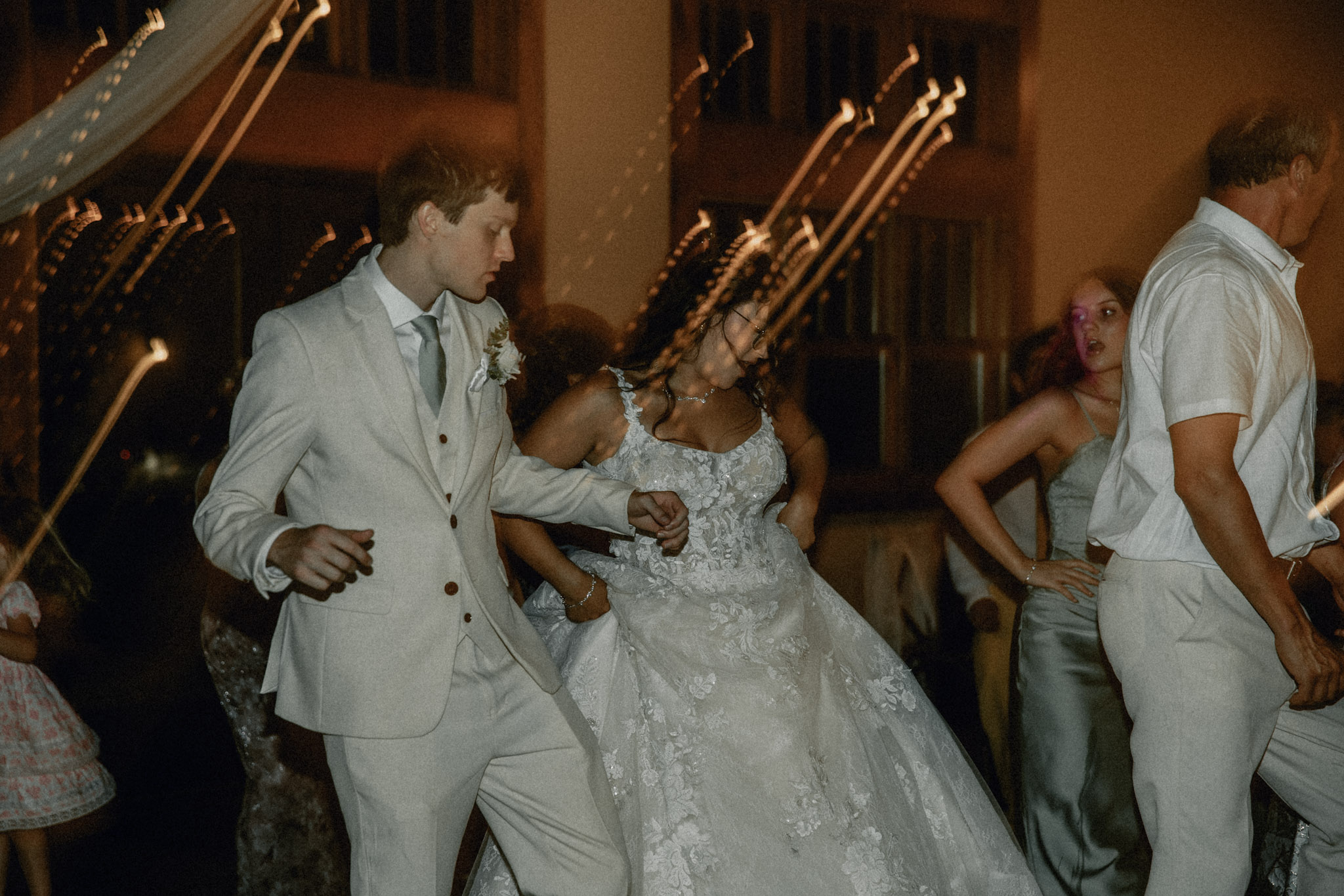 People dancing at a wedding reception, with a woman in a wedding dress and others in formal attire, in a decorated indoor venue.