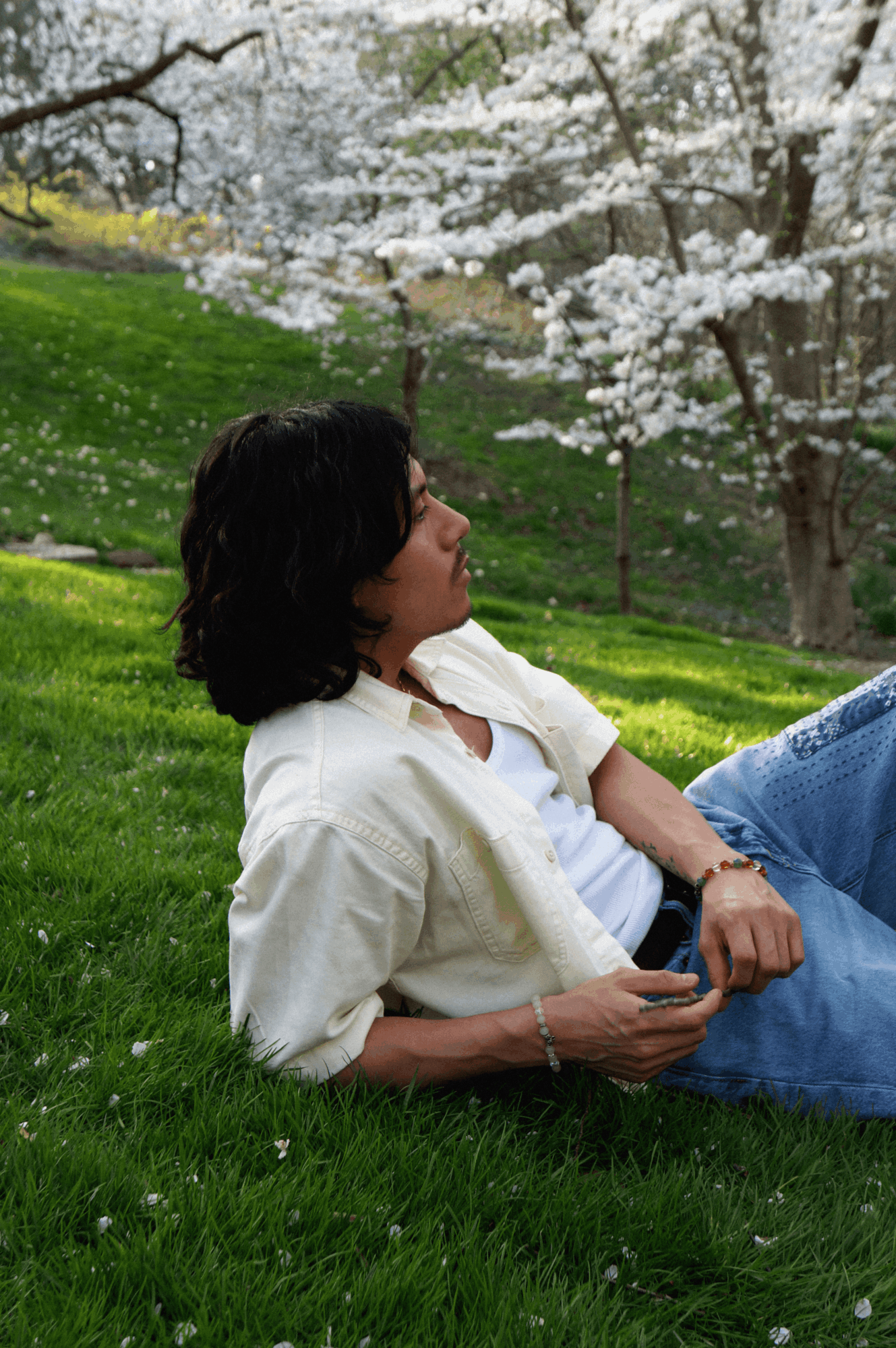 Woman with dark hair lying on grass near blooming trees, wearing a white shirt and jeans, holding a phone.