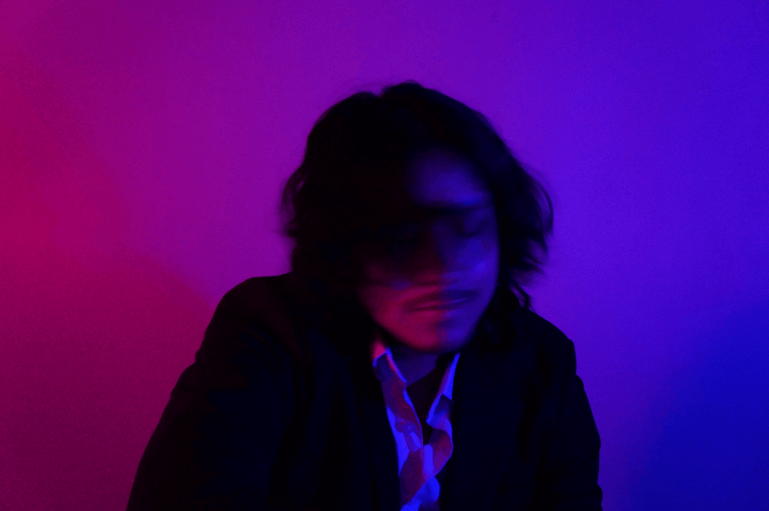 Person with shoulder-length hair in a dark jacket, illuminated by purple and blue lighting, looking downward.