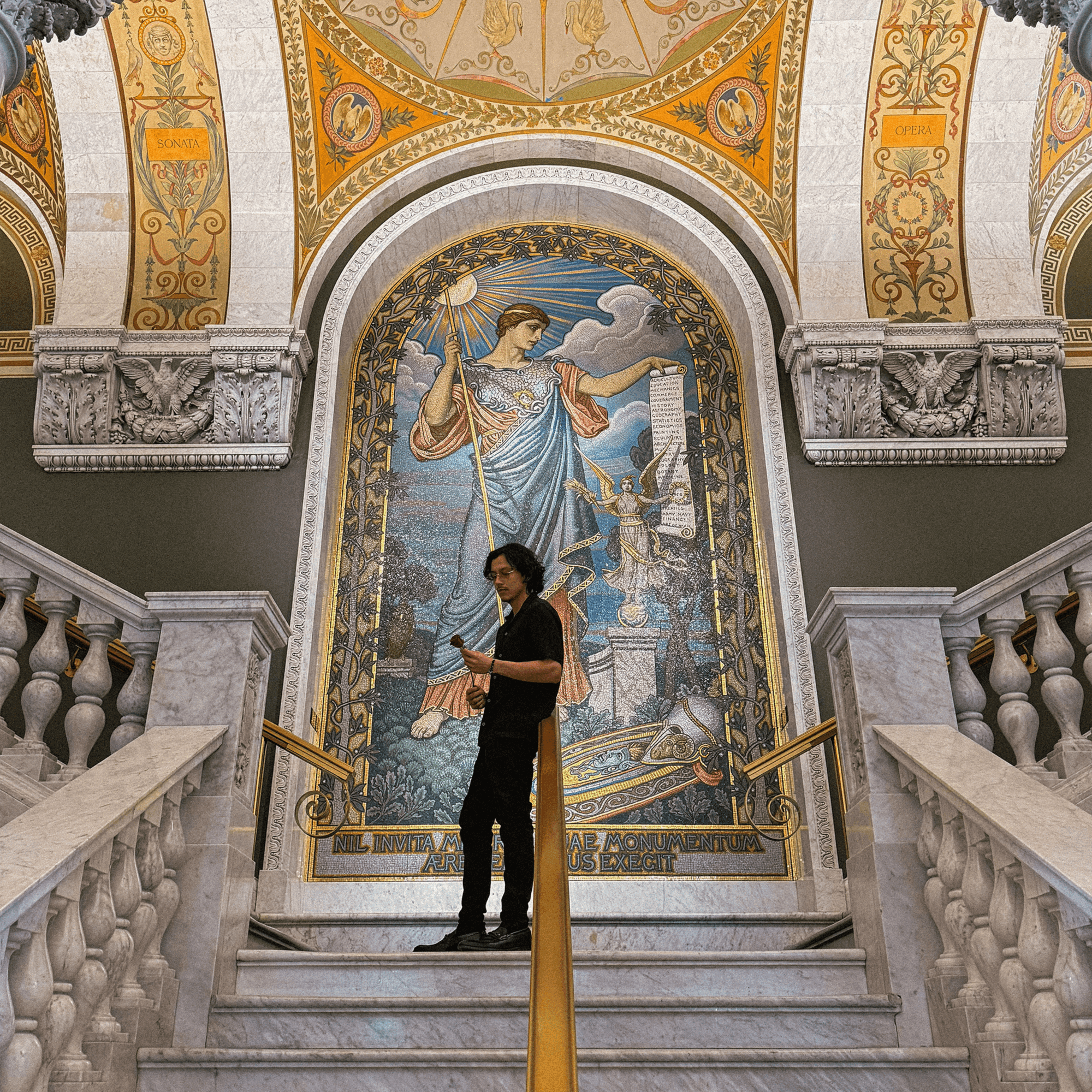 Person standing on stairs in front of a large, ornate mosaic depicting a woman with a staff, inside a decorated building.