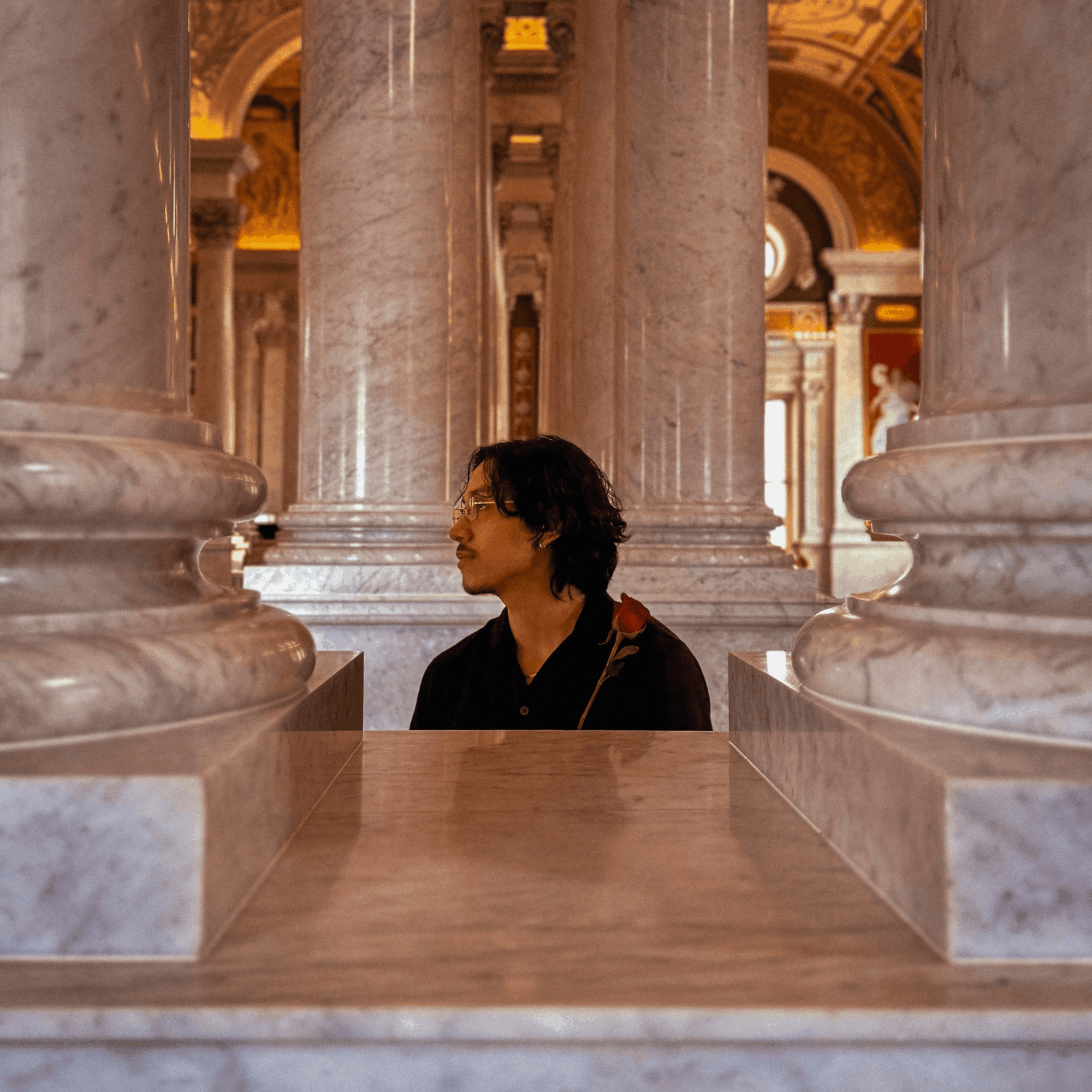 Person with short dark hair and glasses holding a red rose, standing between large marble columns inside a grand building.
