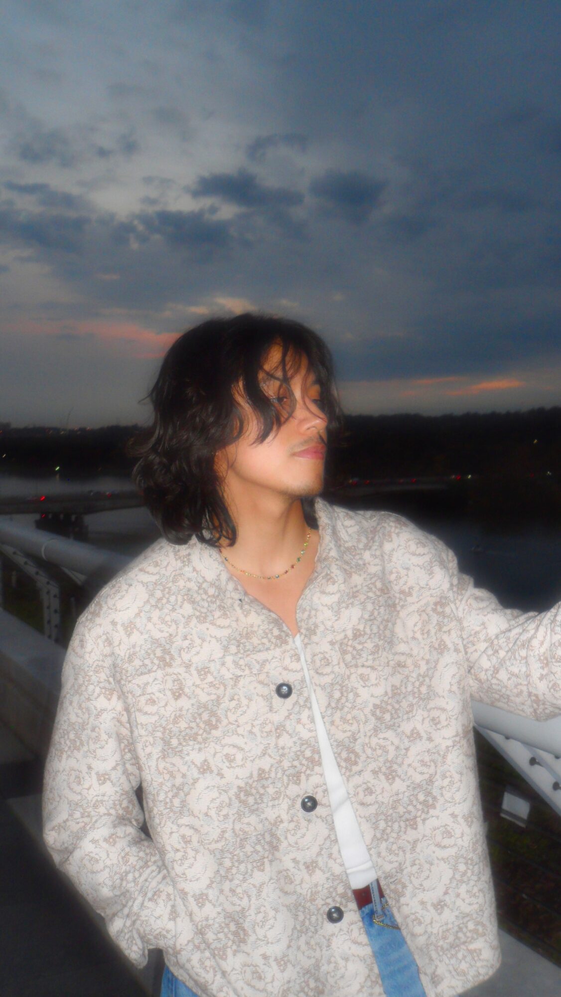 Person with dark, curly hair wearing a patterned jacket outdoors during evening or dusk.