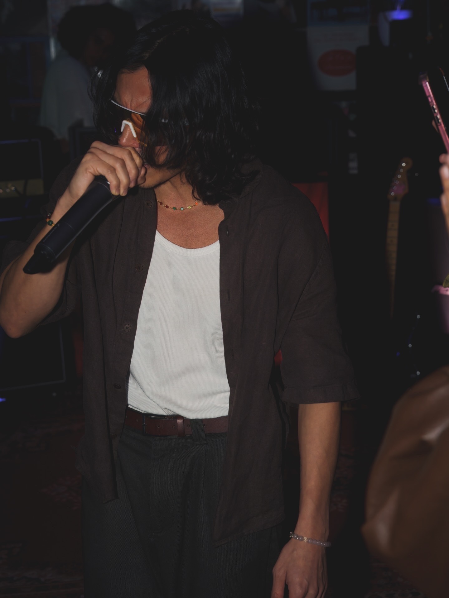 Person with curly hair holding a microphone, wearing a white top and dark jacket, in a dimly lit setting.