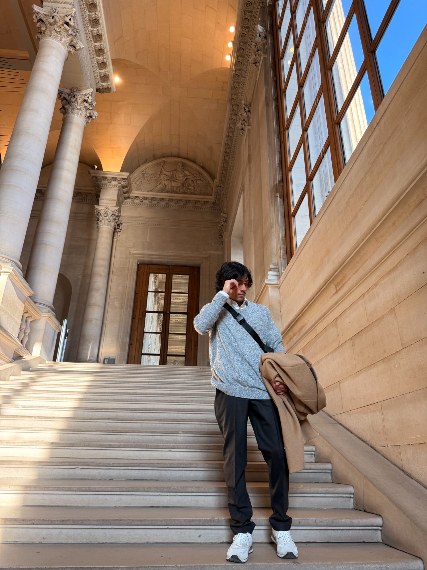 Young man standing on stairs inside a grand building with tall columns and large windows, holding a bag and looking at the camera.