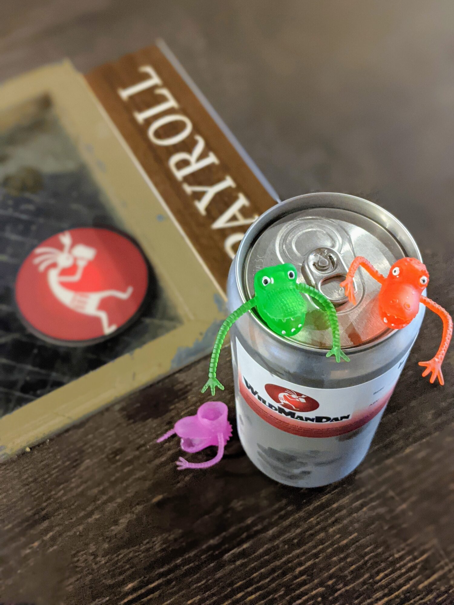 Three small rubber frogs in green, orange, and purple sit on a soda can, with a book and a sticker in the background.
