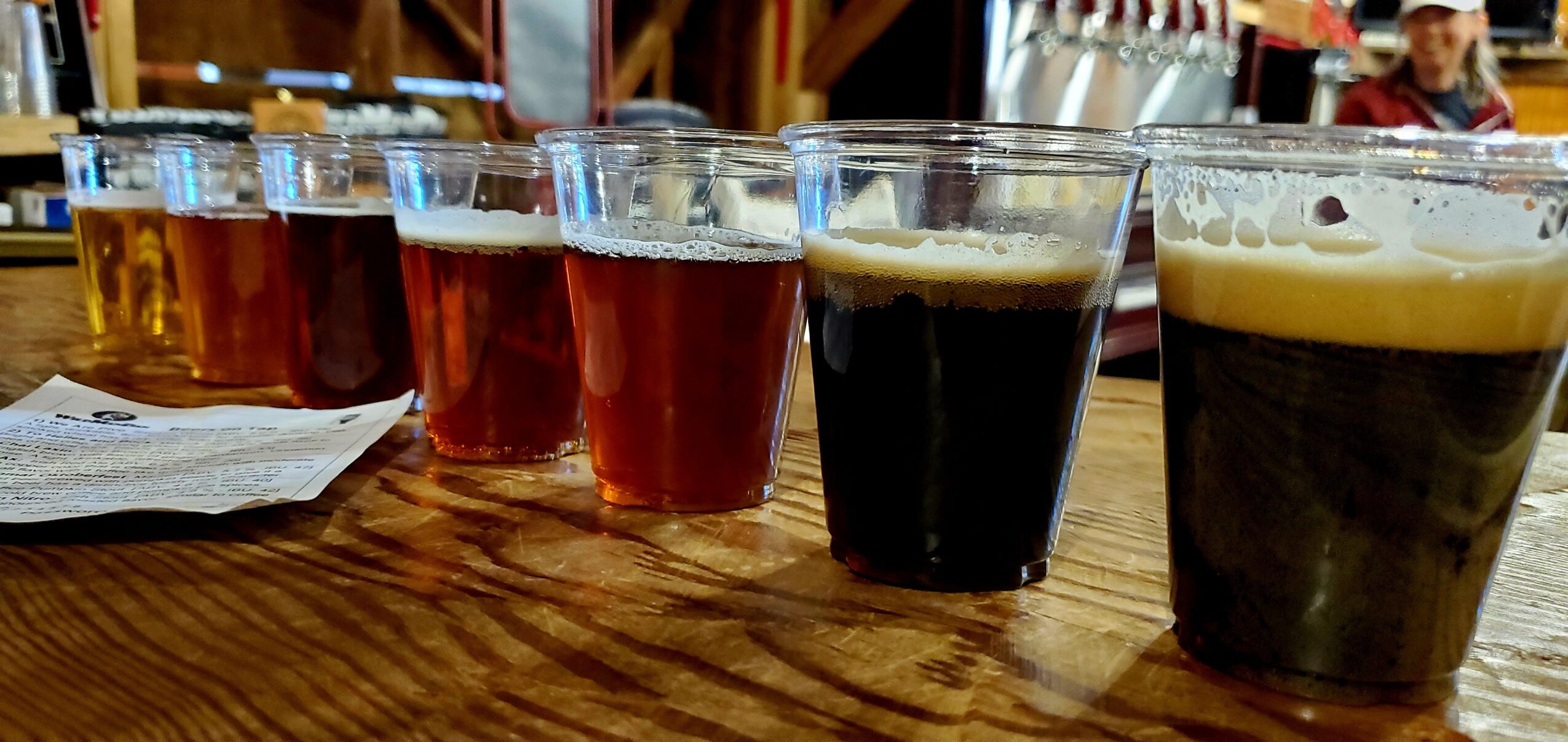 Six glasses of beer on a wooden table, with a bar or brewery setting in the background.
