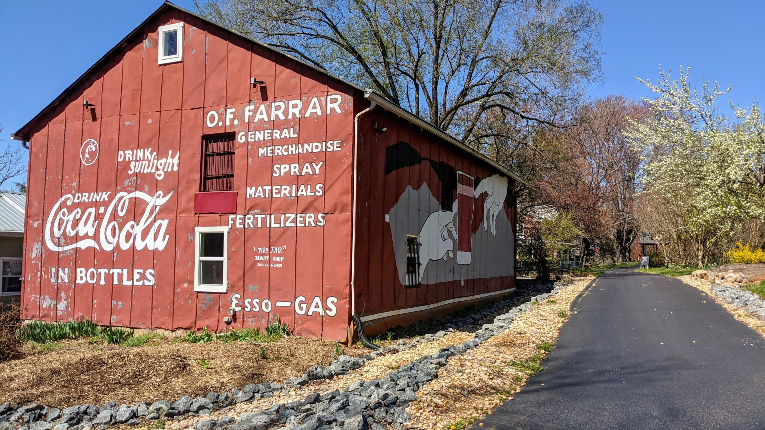 Red barn with white text and logo, next to a paved road and trees, under a clear blue sky.