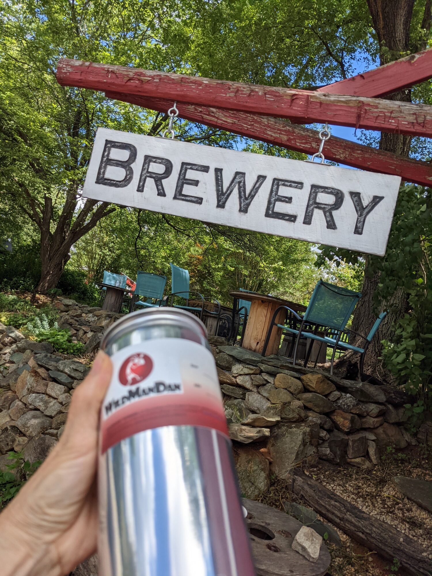 Person holding a can in front of a brewery sign outdoors, with trees and stacked firewood in the background.
