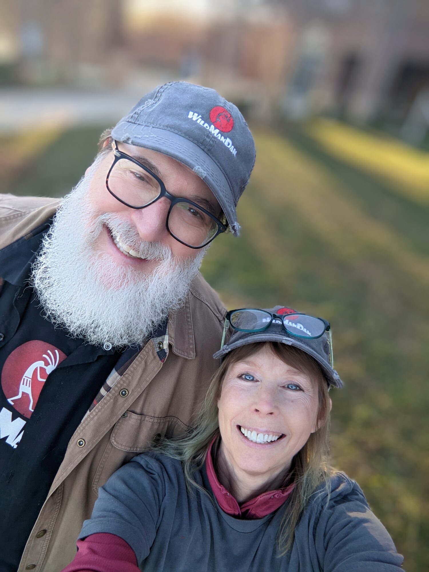 Two smiling people outdoors, one with glasses and a beard, the other with sunglasses on cap, both wearing jackets.