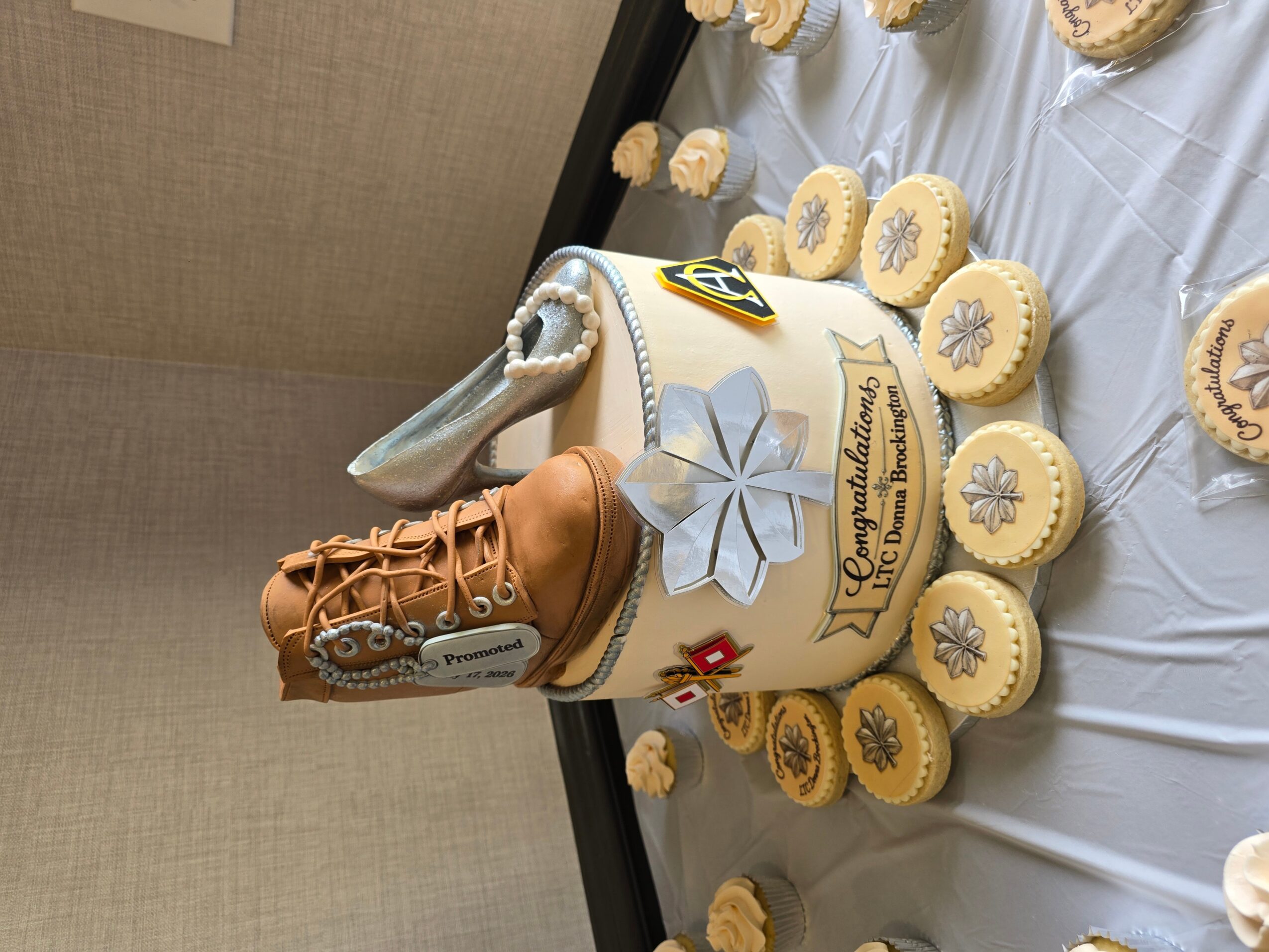 Celebration cake with a boot decoration, surrounded by cookies on a table, with a white ribbon and a blue flower, in a room.