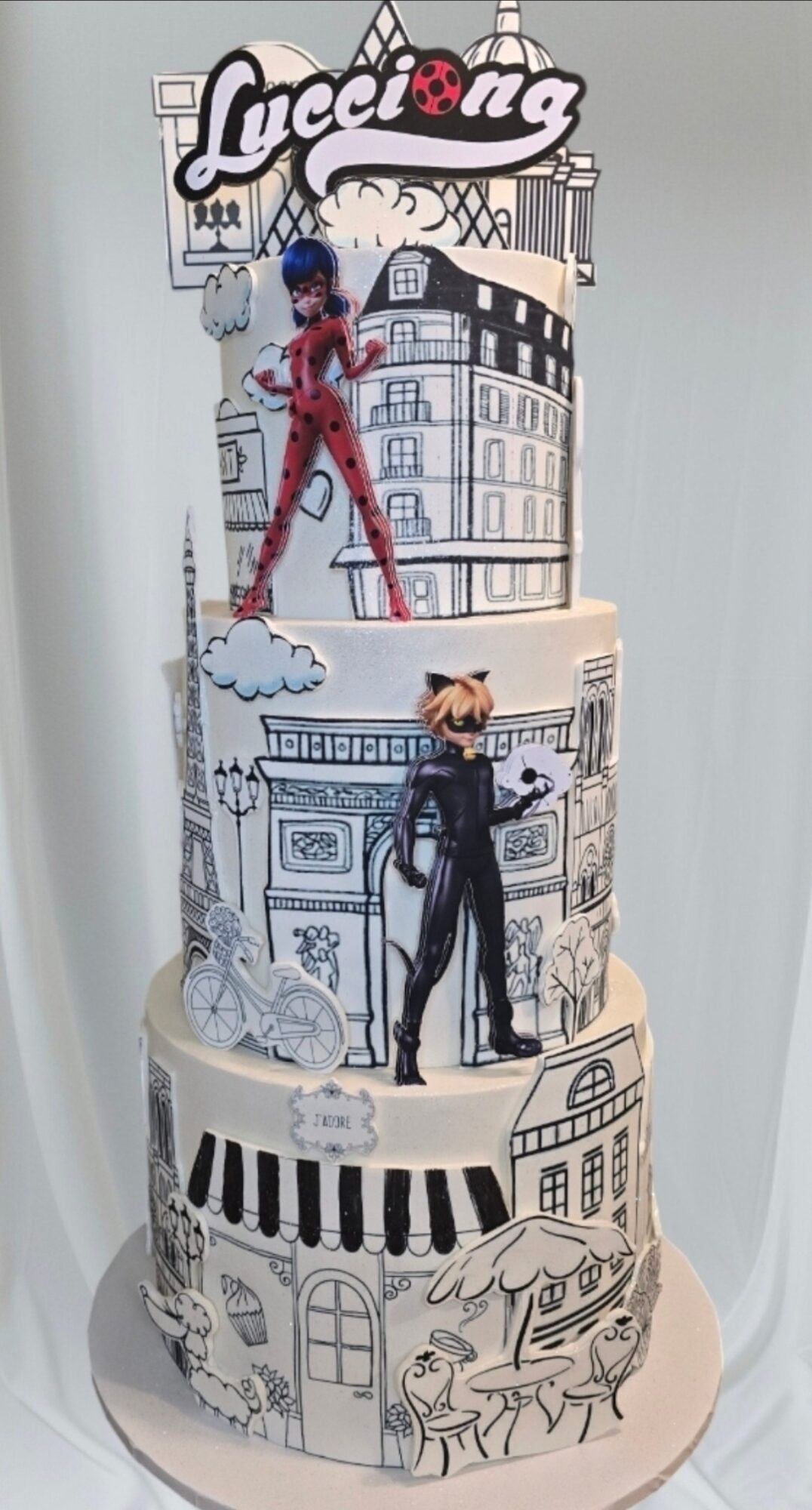 Three-tiered cake with black and white cityscape illustrations, featuring two fashion-themed figurines, and a 'Fashion' sign on top.