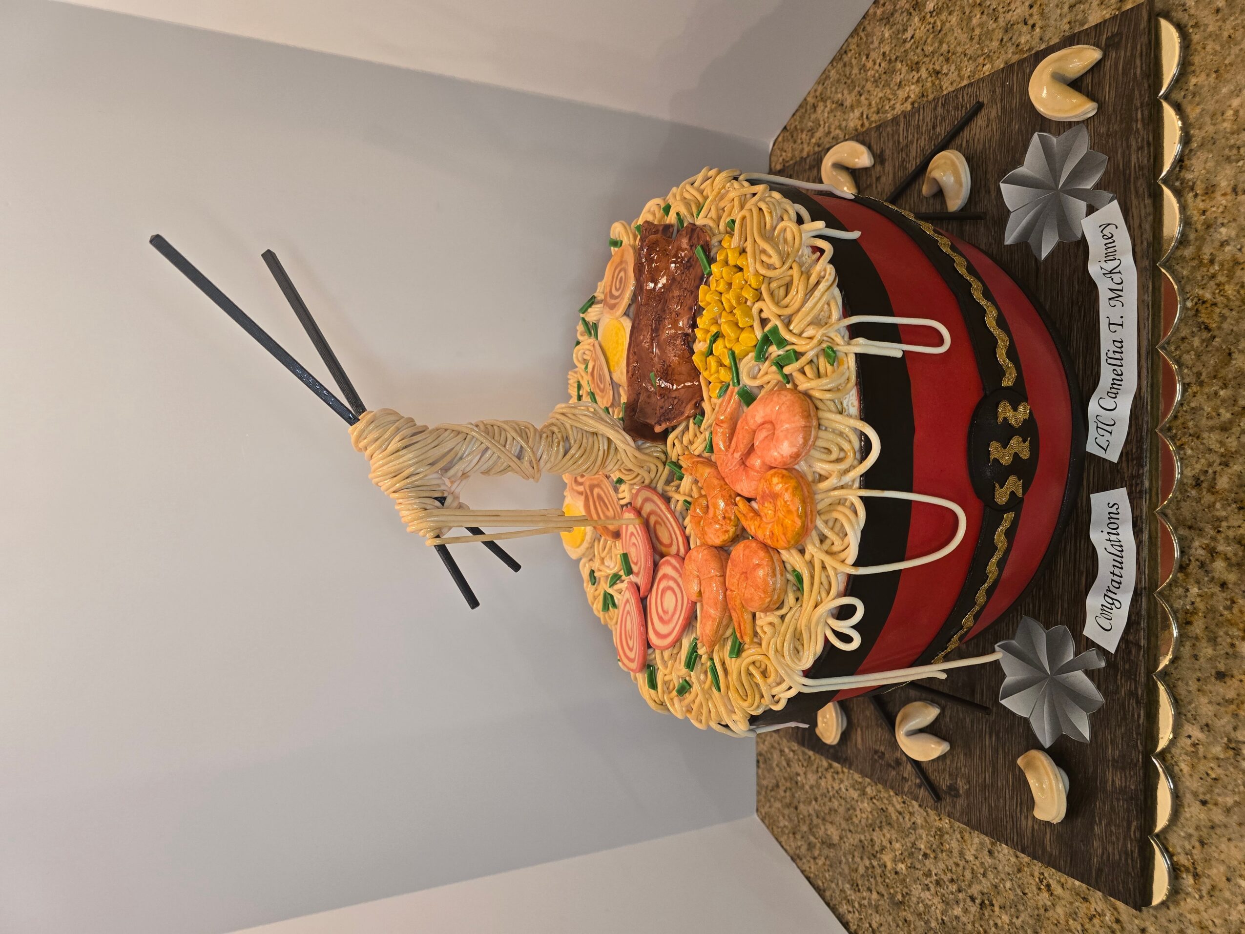 A birthday cake with a noodle tower, shrimp, and decorative elements on top, with chopsticks and a message on the side.
