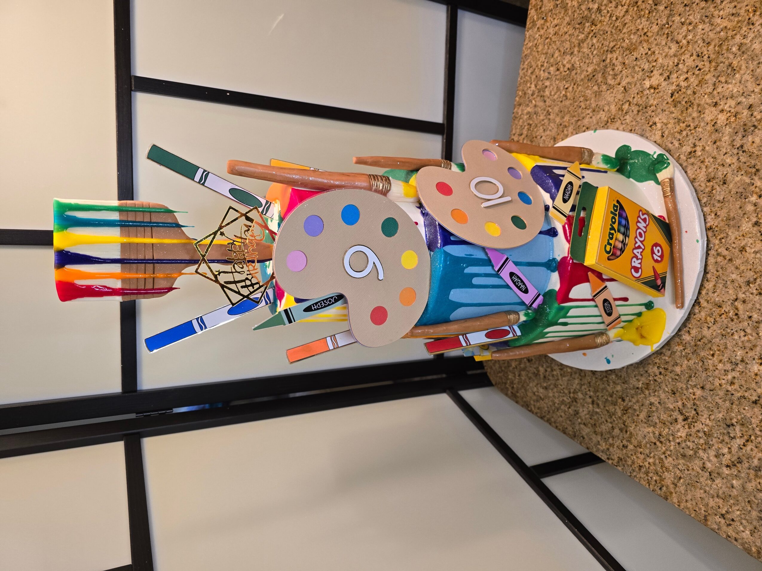Colorful art supplies in a white container, including paint palettes, brushes, and markers, on a speckled surface.