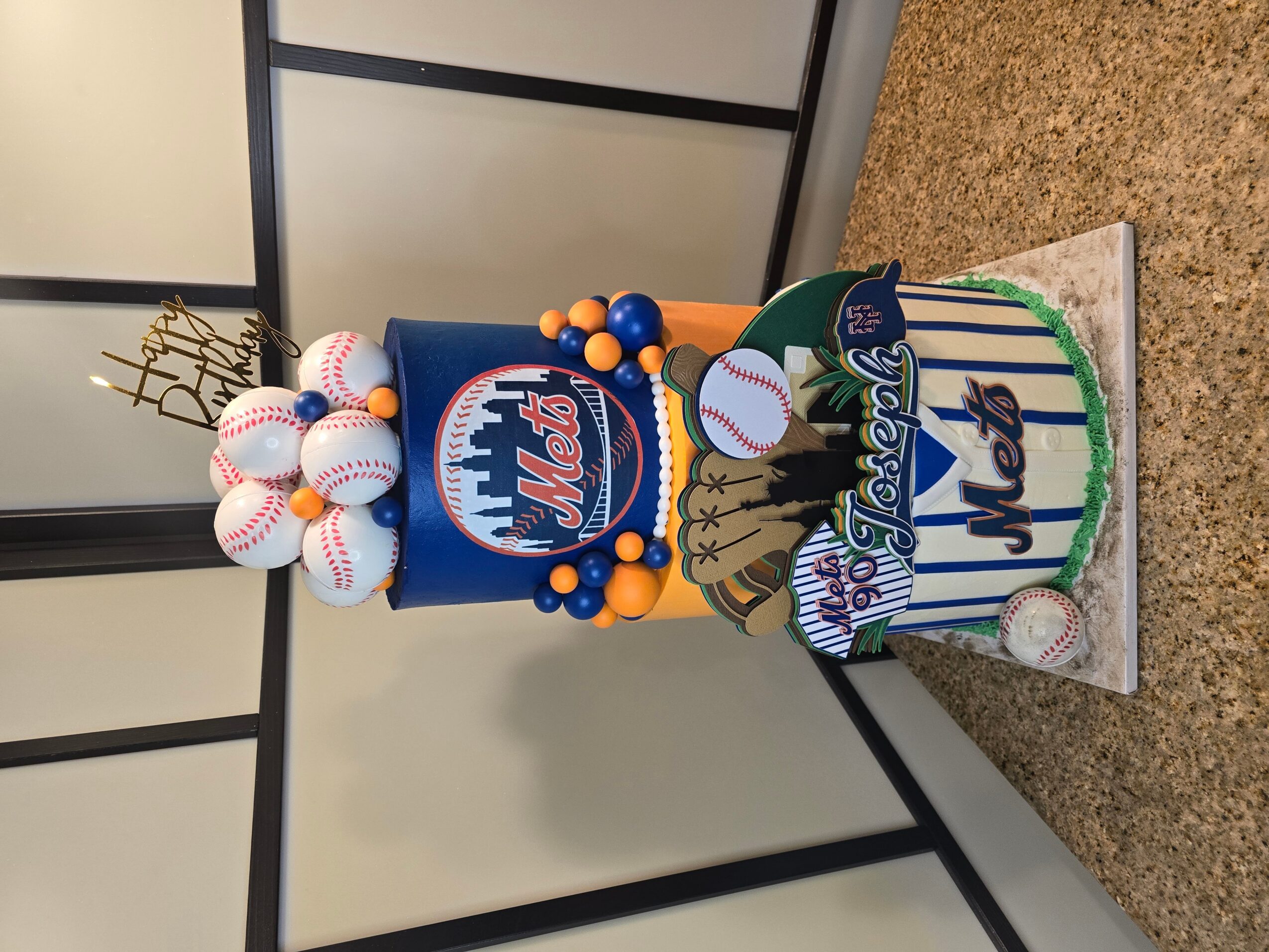 Baseball-themed cake with baseballs, a city skyline, and a baseball glove decoration, celebrating Mets baseball team.