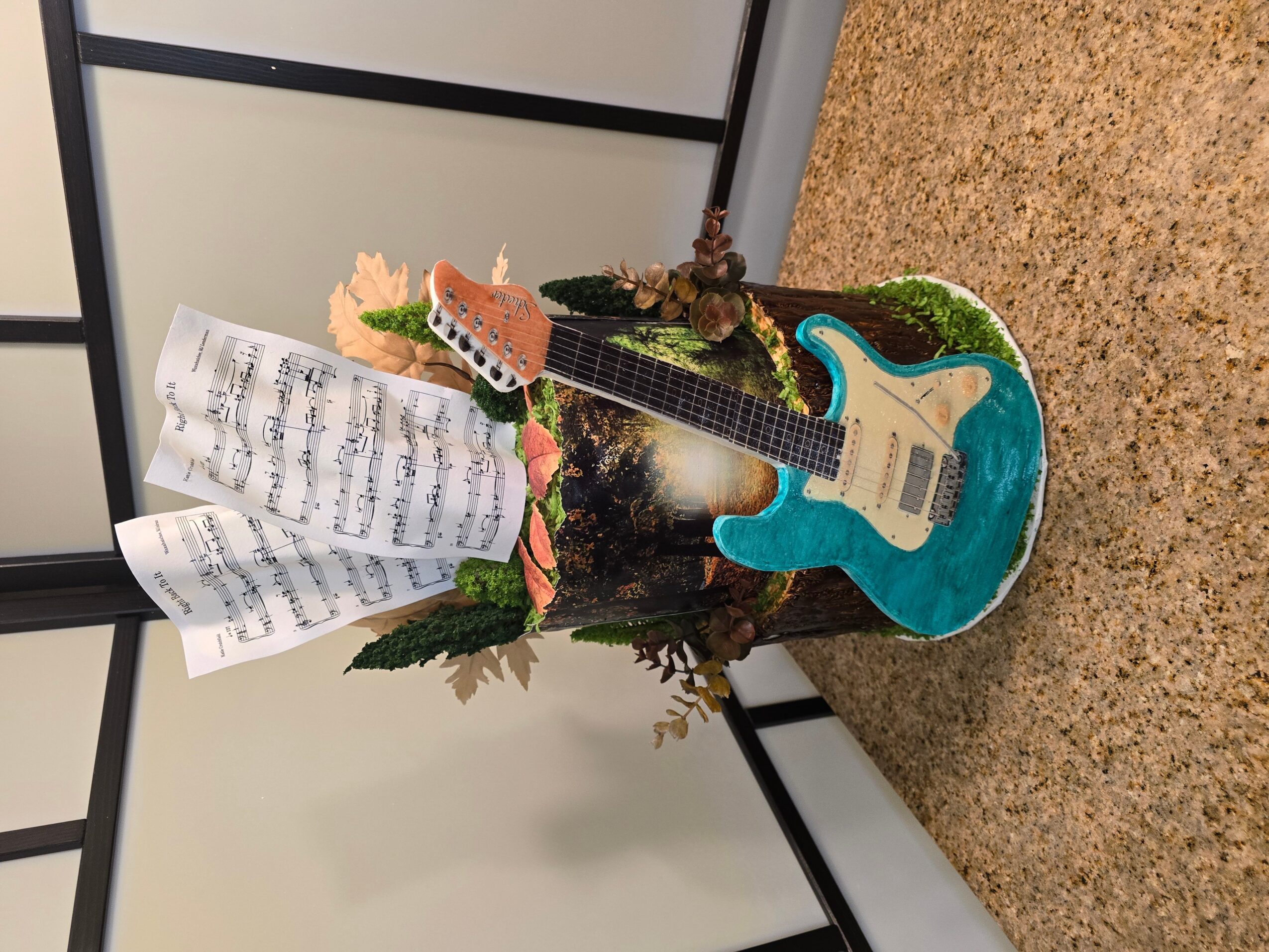 Decorative guitar with sheet music, flowers, and foliage, placed on a beige carpeted floor against a white wall.