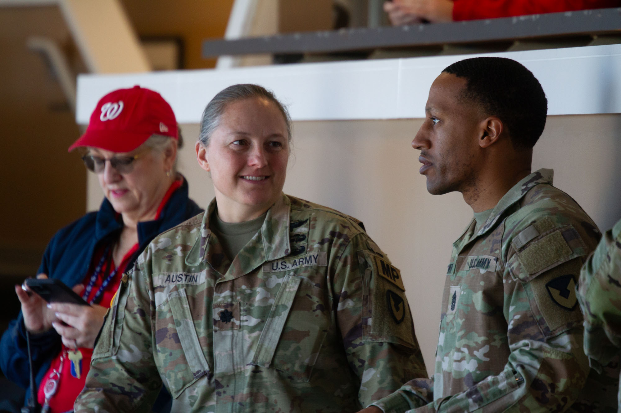 Two military personnel in uniform engaged in conversation, with a woman in casual clothing and a red cap nearby, in an indoor setting.