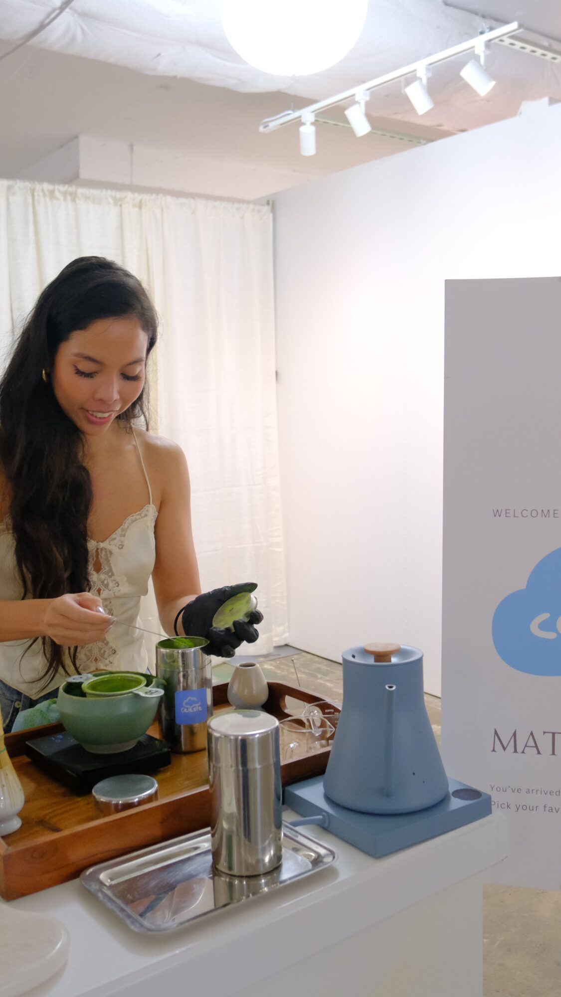 Woman with long dark hair preparing food in a kitchen, with a blue kettle and various utensils on the counter.