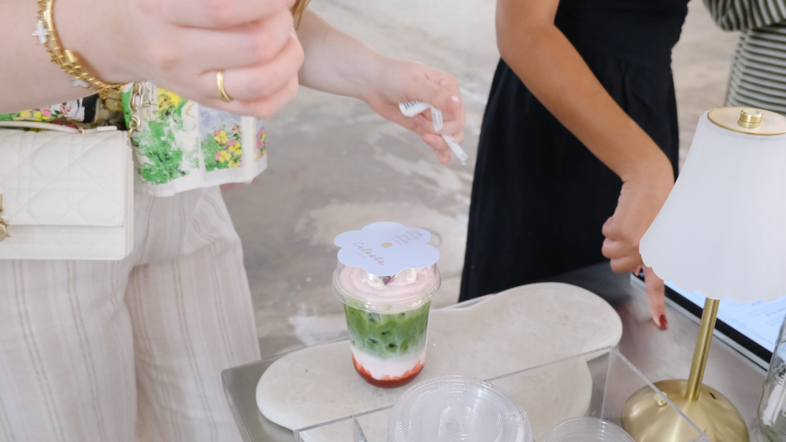 People gather around a table with a layered dessert in a glass, with hands reaching towards it, and a lamp nearby.