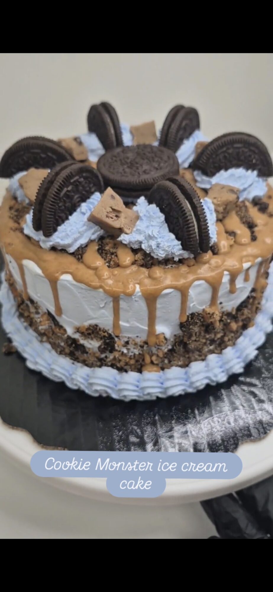 Cake decorated with Oreo cookies, blue frosting, and caramel, with a white base and drizzled caramel topping.