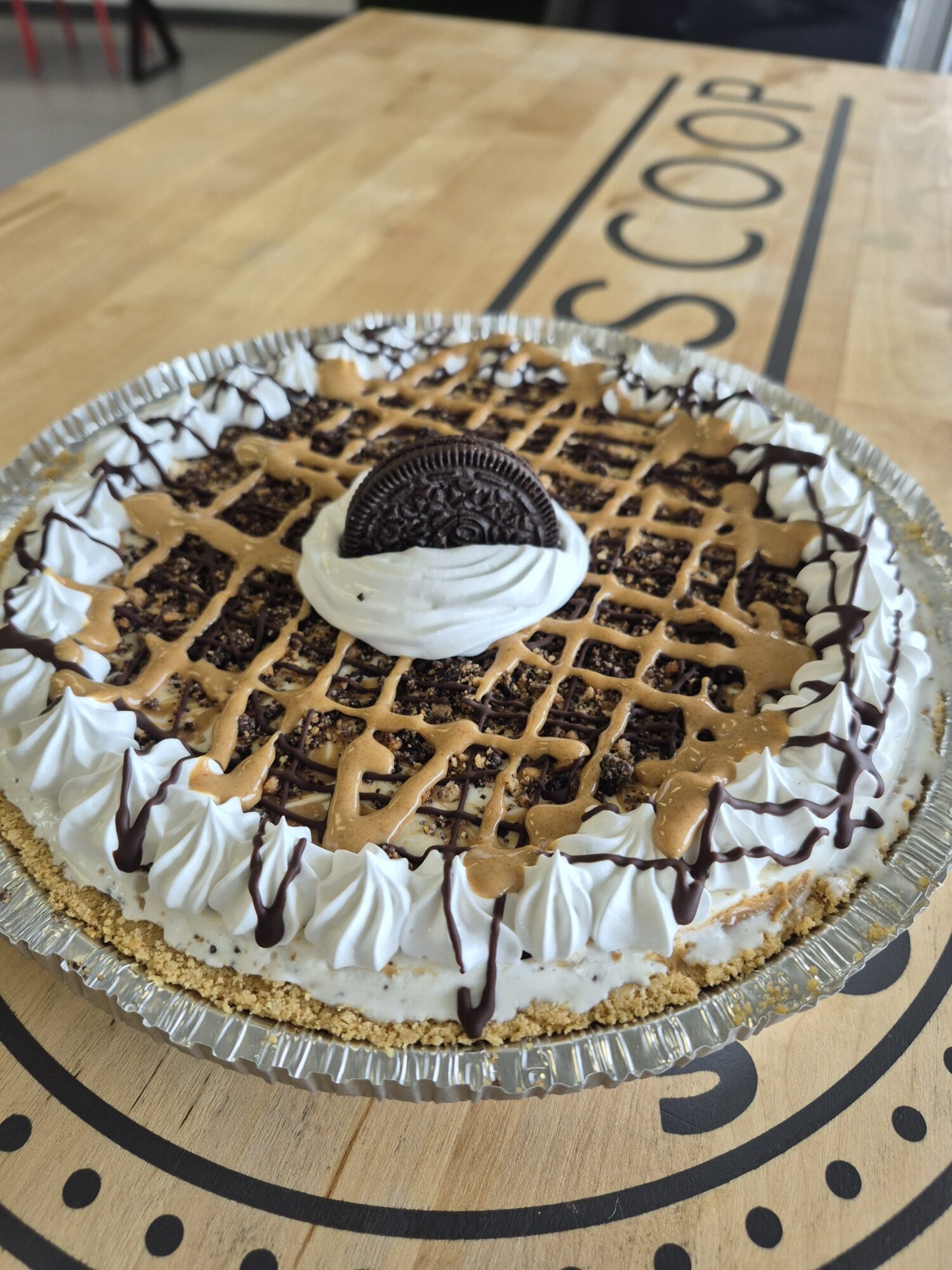 A round dessert with whipped cream, chocolate drizzle, and an Oreo cookie on top, on a table with a logo.