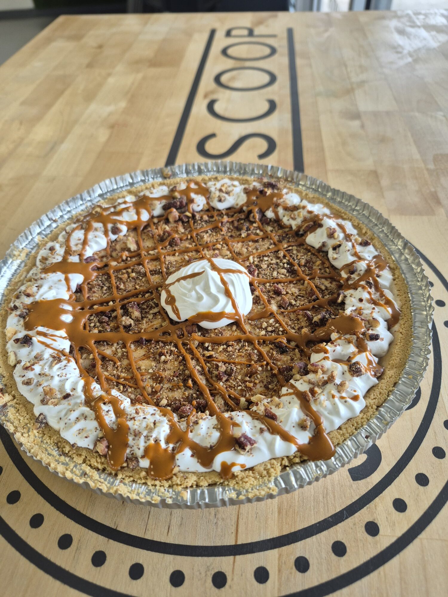 Pie with whipped cream, caramel drizzle, and crumb topping on a silver tray, on a wooden table with 'SCOOP' text.
