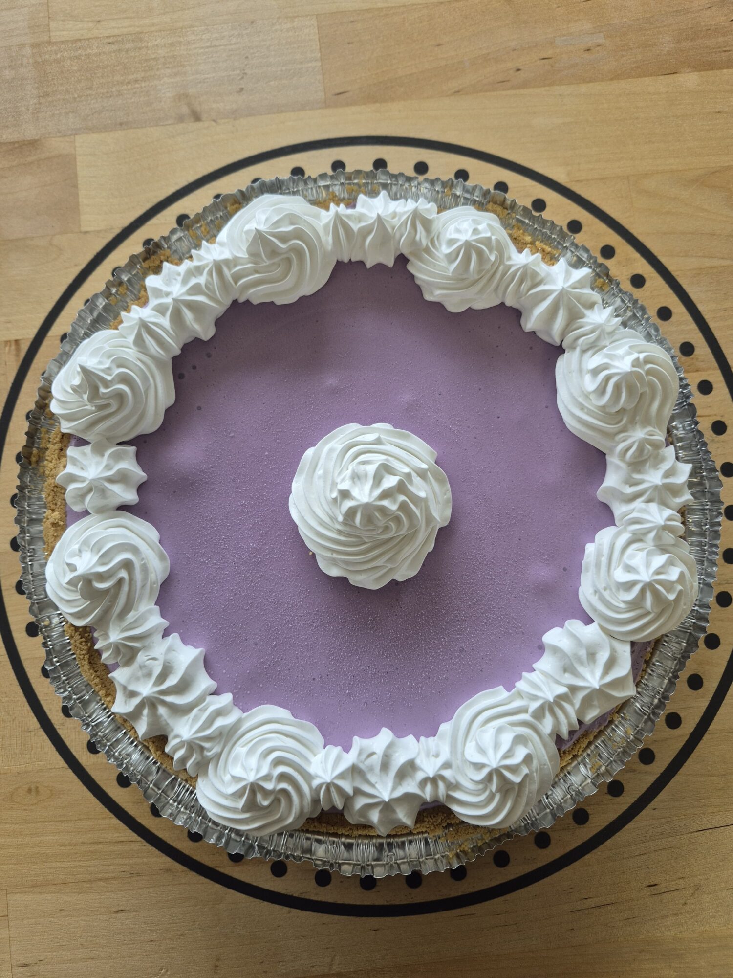 Round cake with purple frosting, decorated with white whipped cream rosettes around the edge and a central rosette, on a decorative plate.