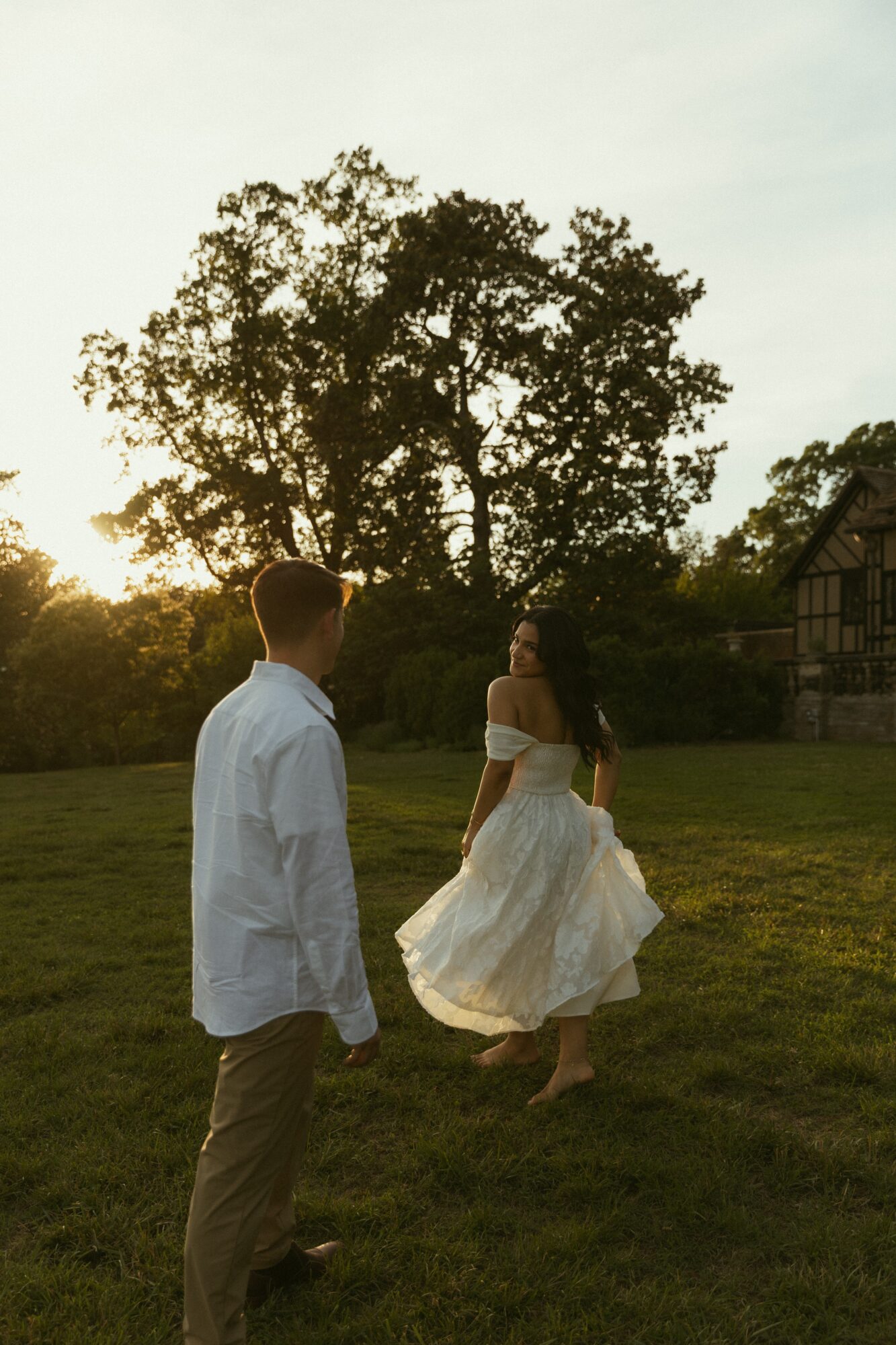 A man and woman stand outdoors on grass, with trees and a building in the background, during daytime.