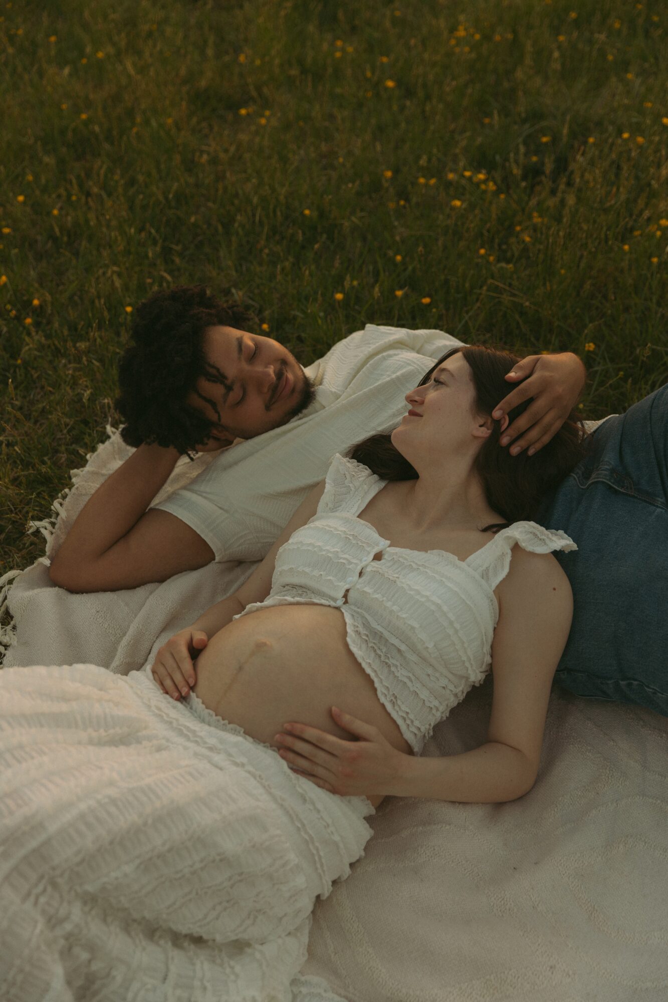 Two people lying on grass, one with a pregnant belly, embracing and looking at each other, surrounded by yellow flowers.