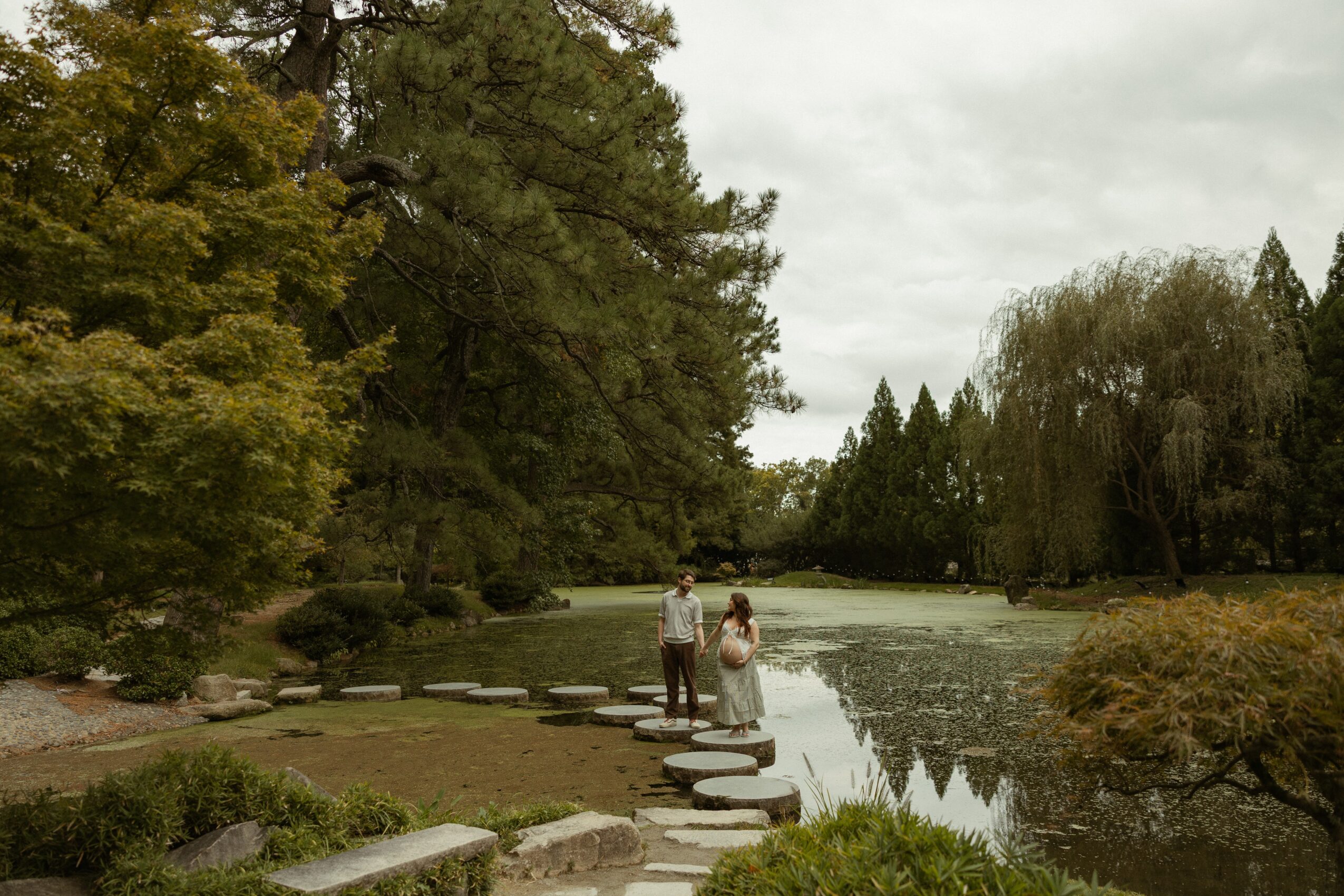 People standing on stepping stones in a river surrounded by trees and greenery, overcast sky, peaceful outdoor scene.