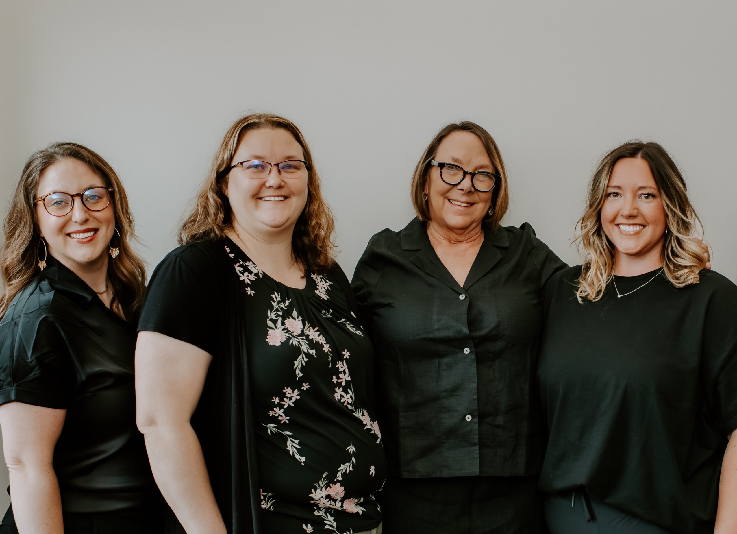 Four women standing side by side, smiling, against a plain light gray background.