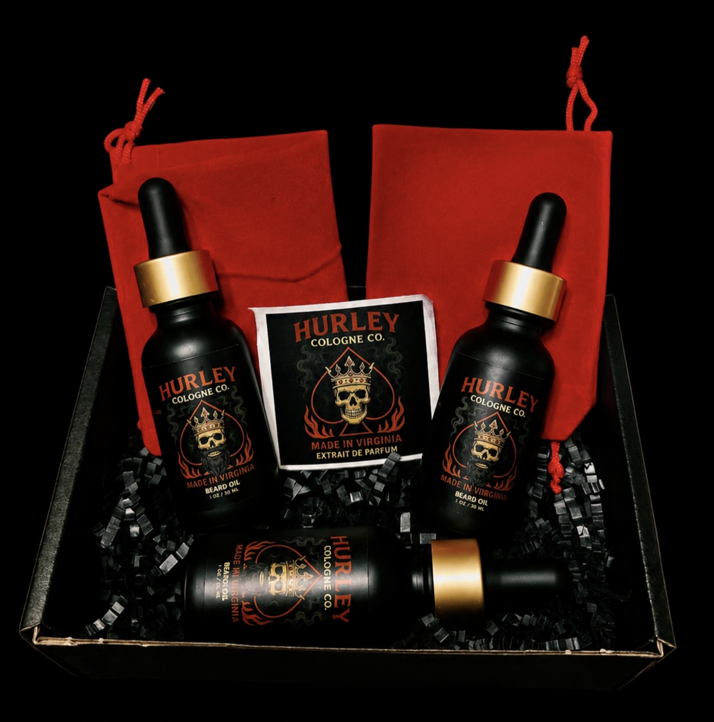 Black box containing three Hurley Cologne bottles with gold caps, two red pouches, and a card with a skull logo, all on black shredded paper.