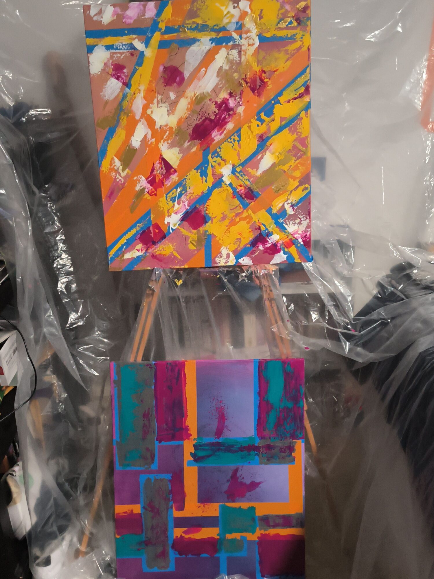 Two colorful abstract paintings on small canvases with wooden sticks, placed on plastic wrapping.