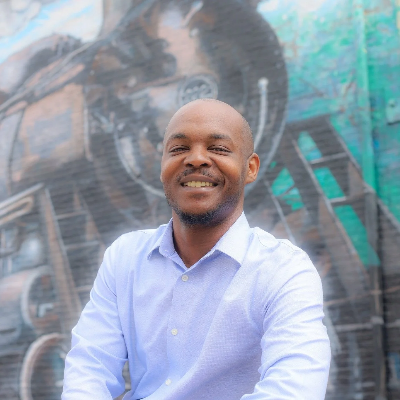 Smiling man in a light blue shirt standing in front of a colorful graffiti wall.