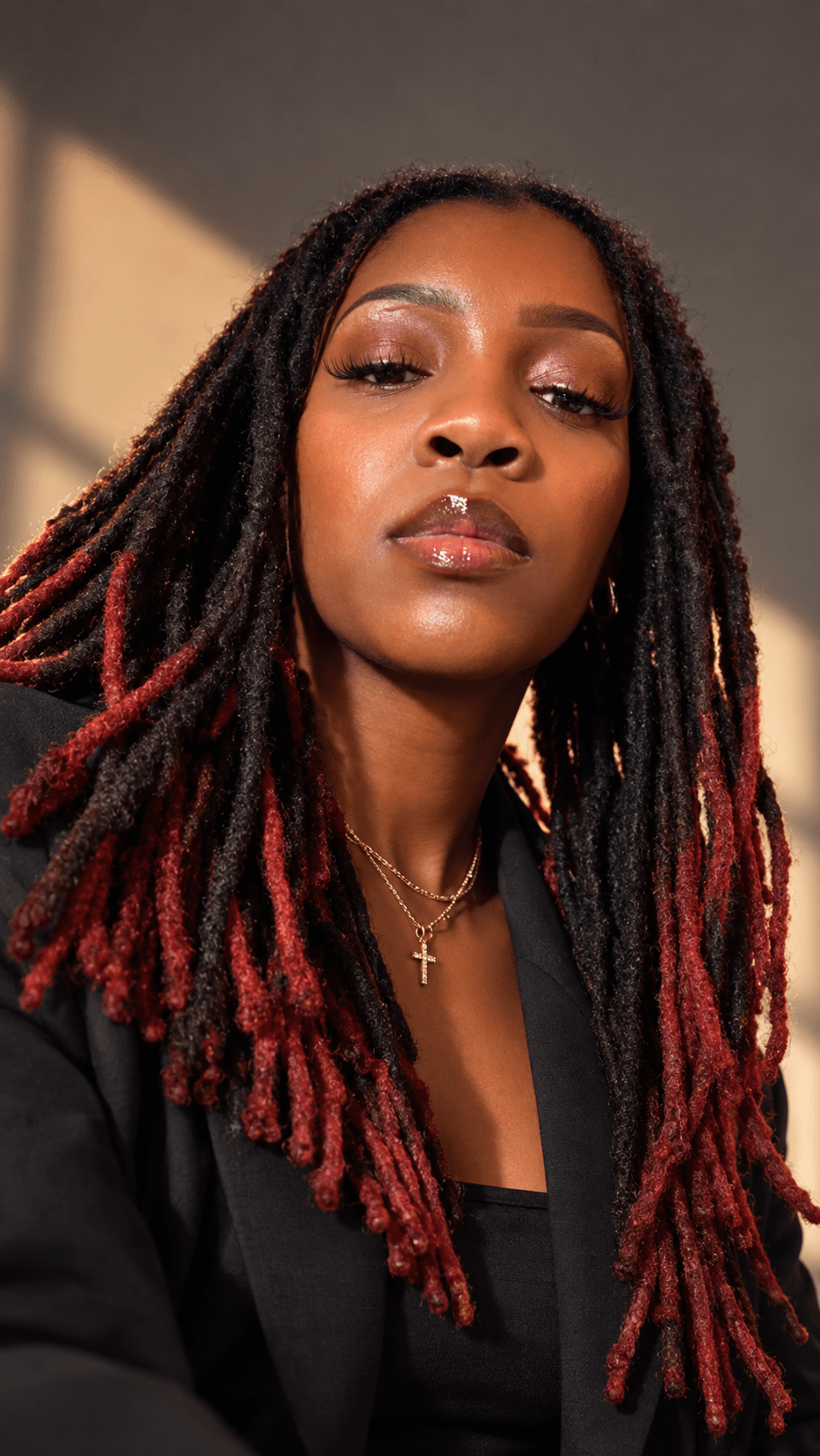 Portrait of a woman with long dreadlocks, wearing a black blazer and a cross necklace, looking at the camera.