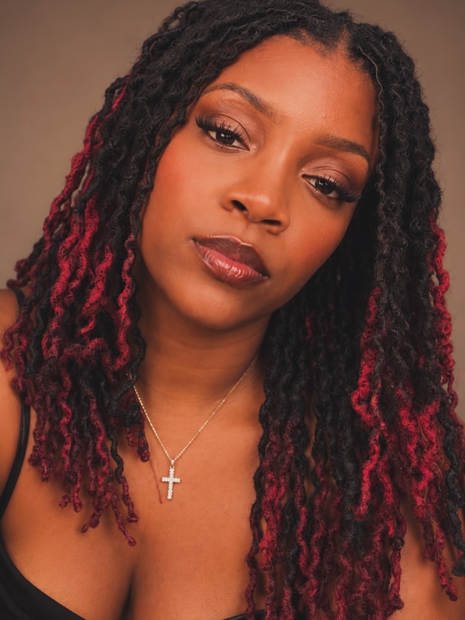 Close-up of a woman with styled hair, wearing makeup and a cross necklace, looking at the camera.