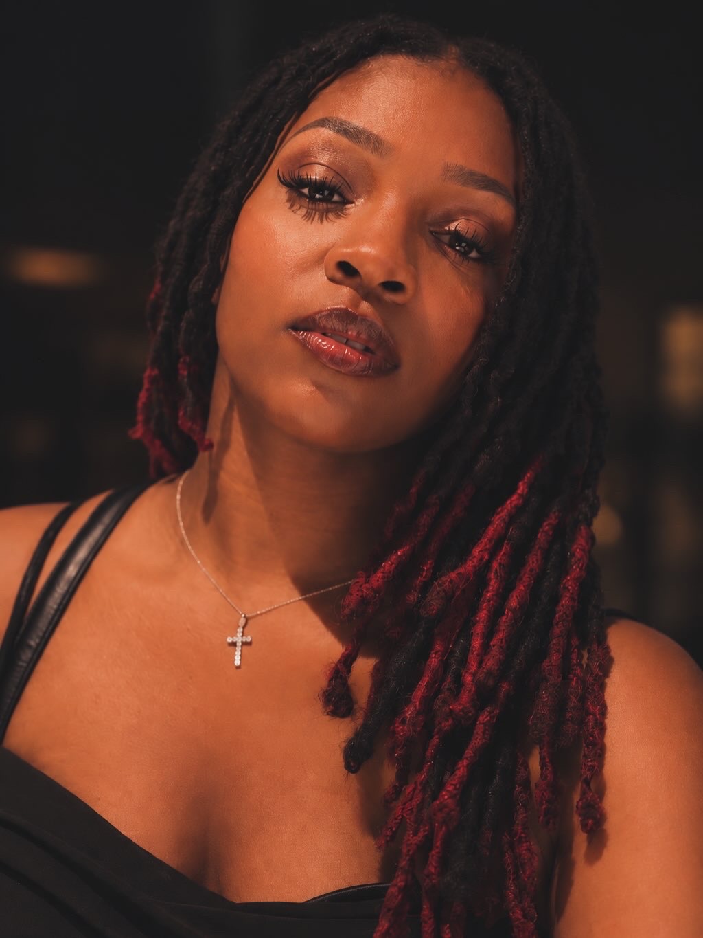 A woman with long dreadlocks, wearing a black top and a cross necklace, looks directly at the camera.
