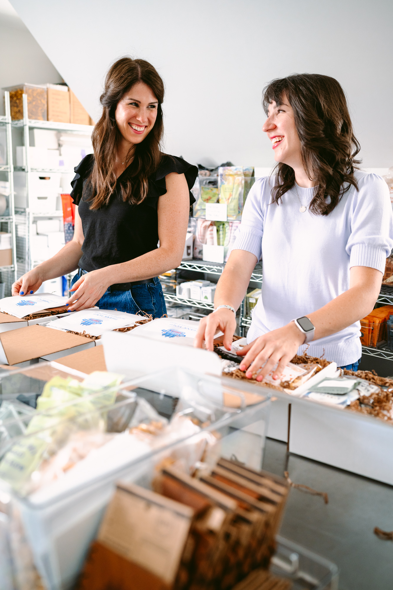 Two women behind a counter in a store, smiling and talking, with various products on the counter.