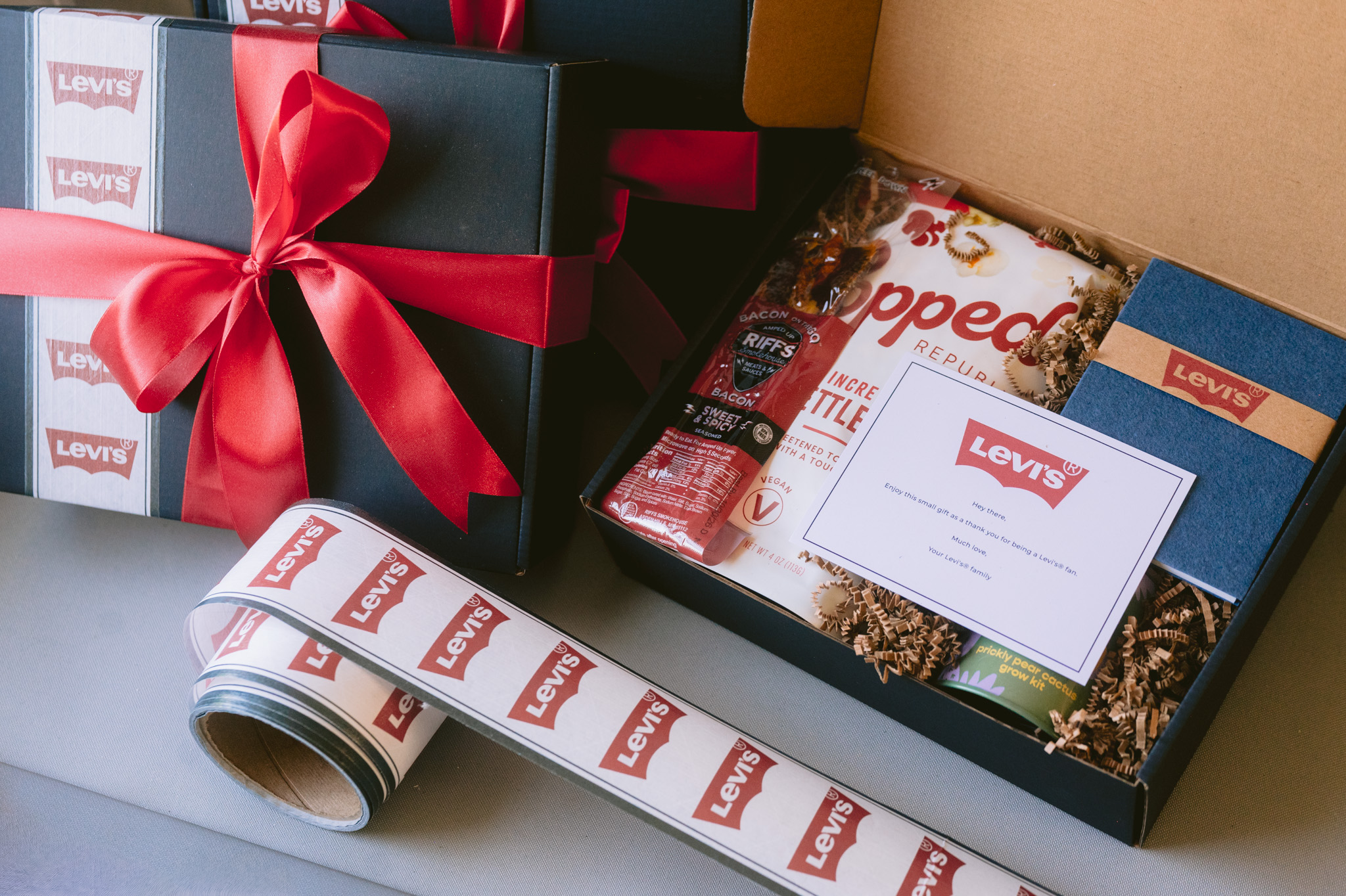 Open gift box with snacks, a card, and a wrapped present with a red ribbon, on a white surface.