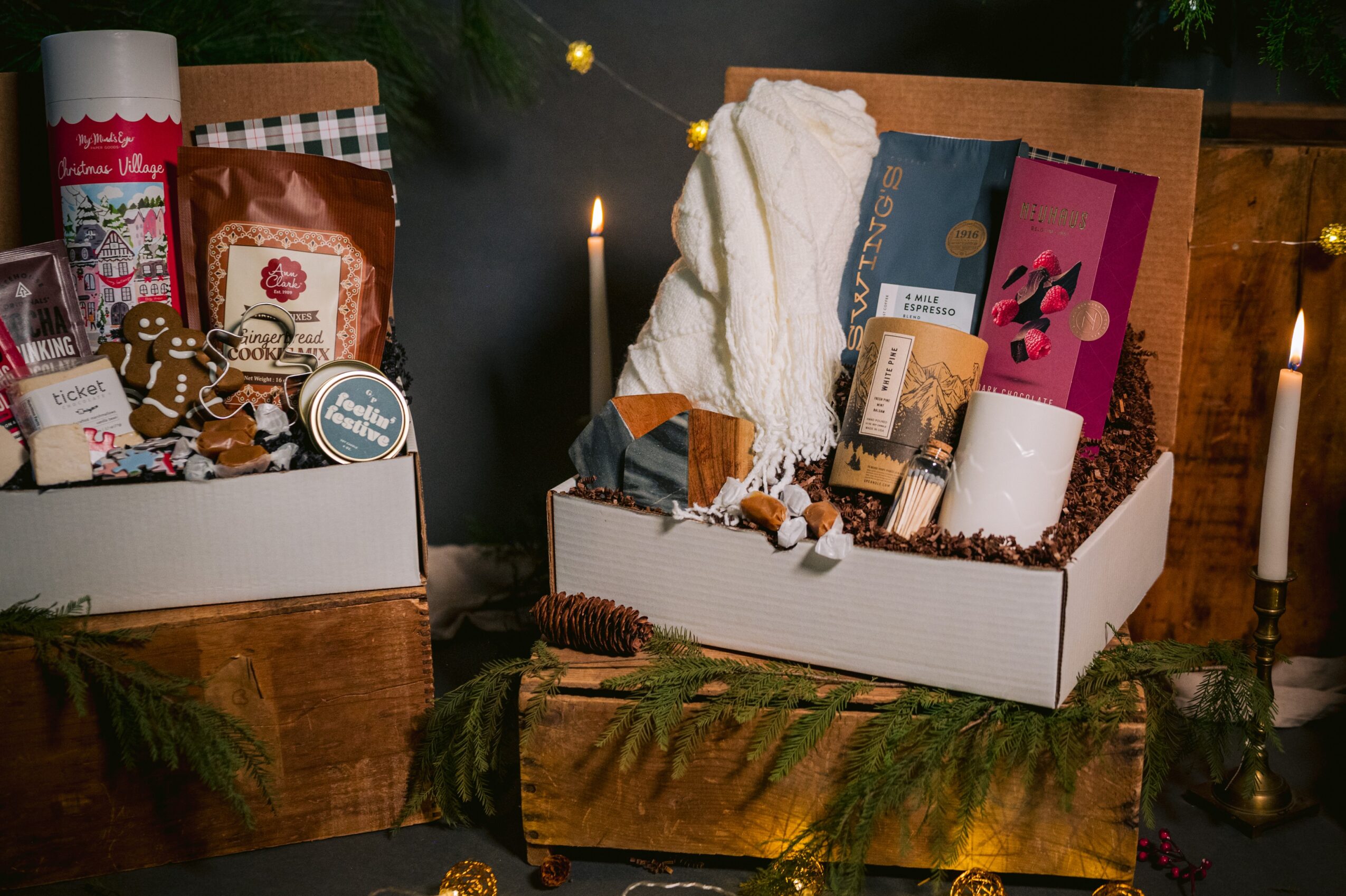 Two boxes filled with gifts, candles, and decorative items on a wooden surface with candles and greenery nearby.