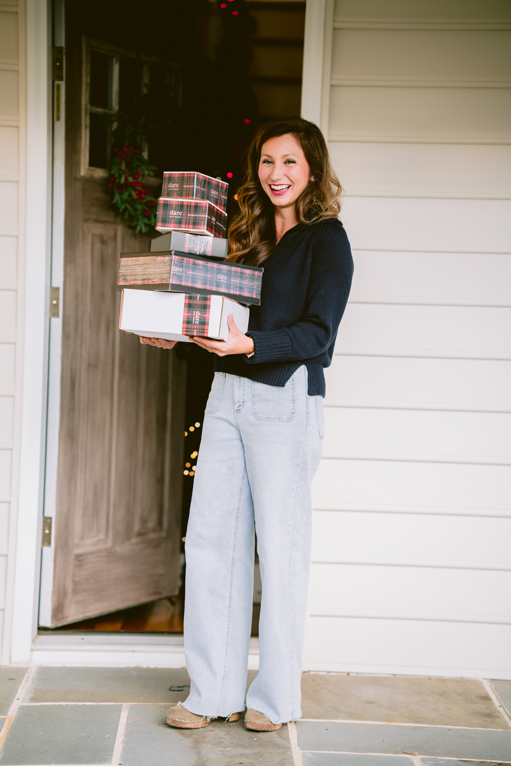 Woman standing at door holding wrapped gifts, smiling, outside house with white siding, holiday decorations visible inside.