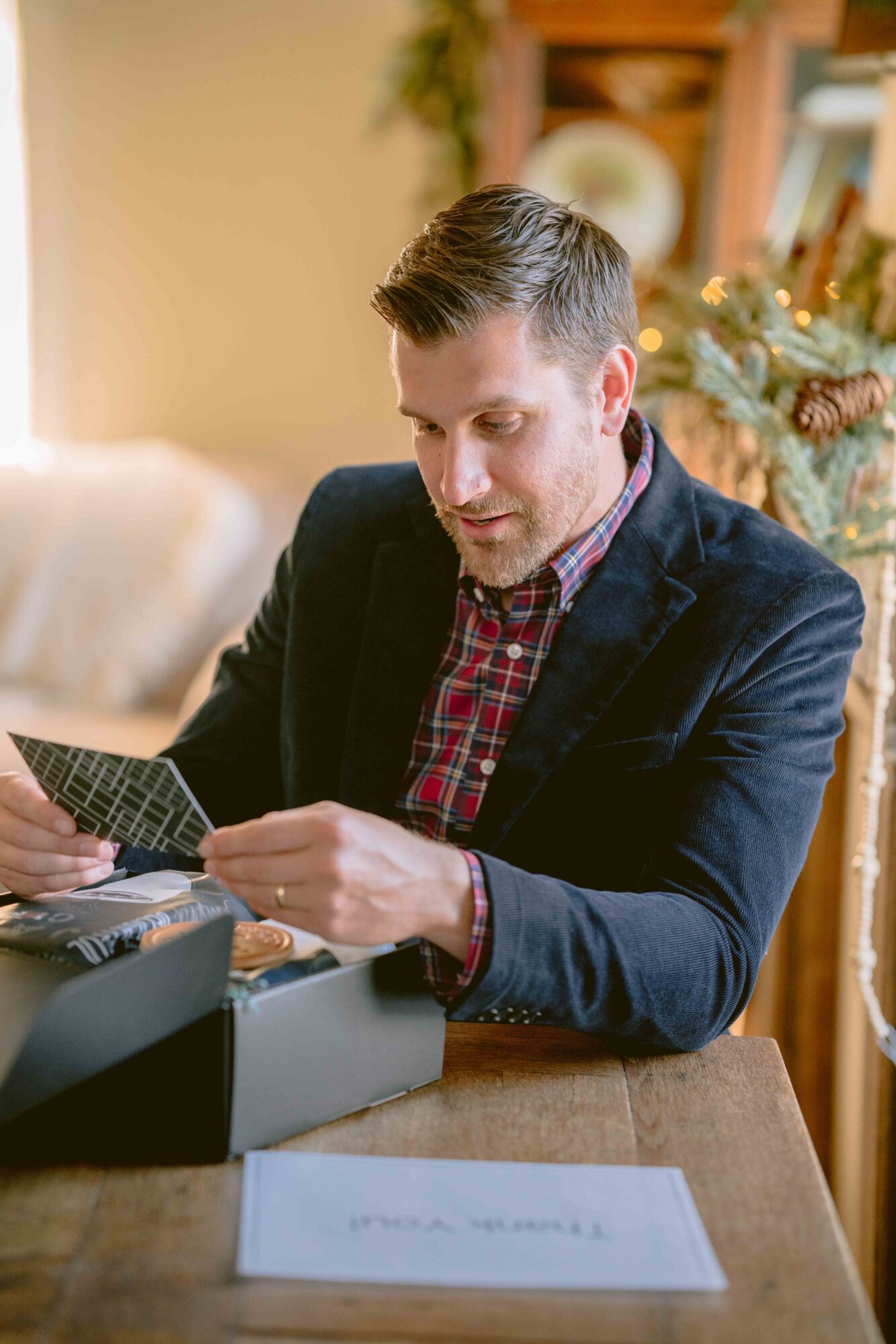 Man in a dark blazer and red checkered shirt looking at photographs, sitting at a table in a cozy room.