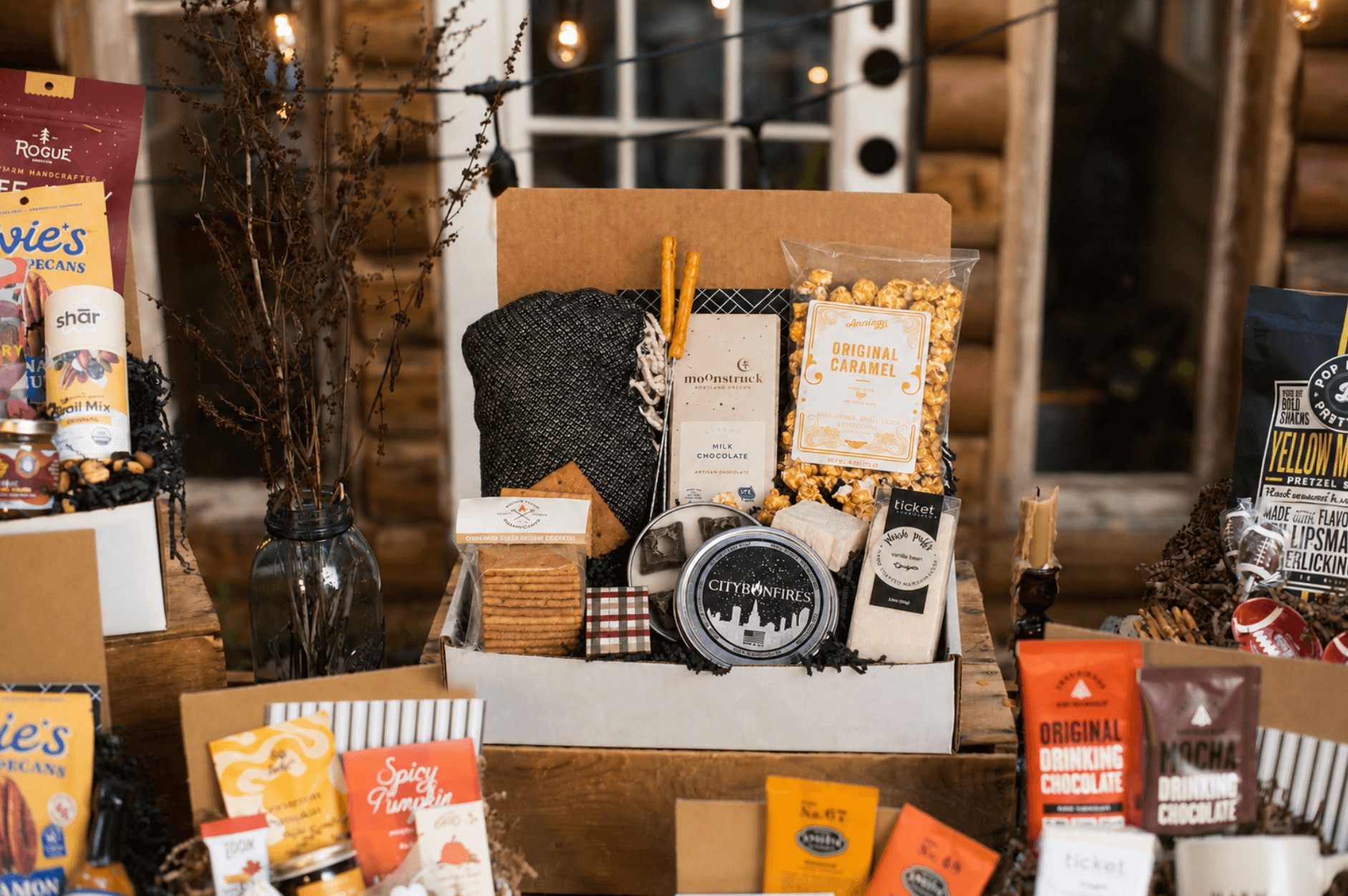 Display of various gift items and snacks on a table with a wooden background, including a framed picture and a black bag.