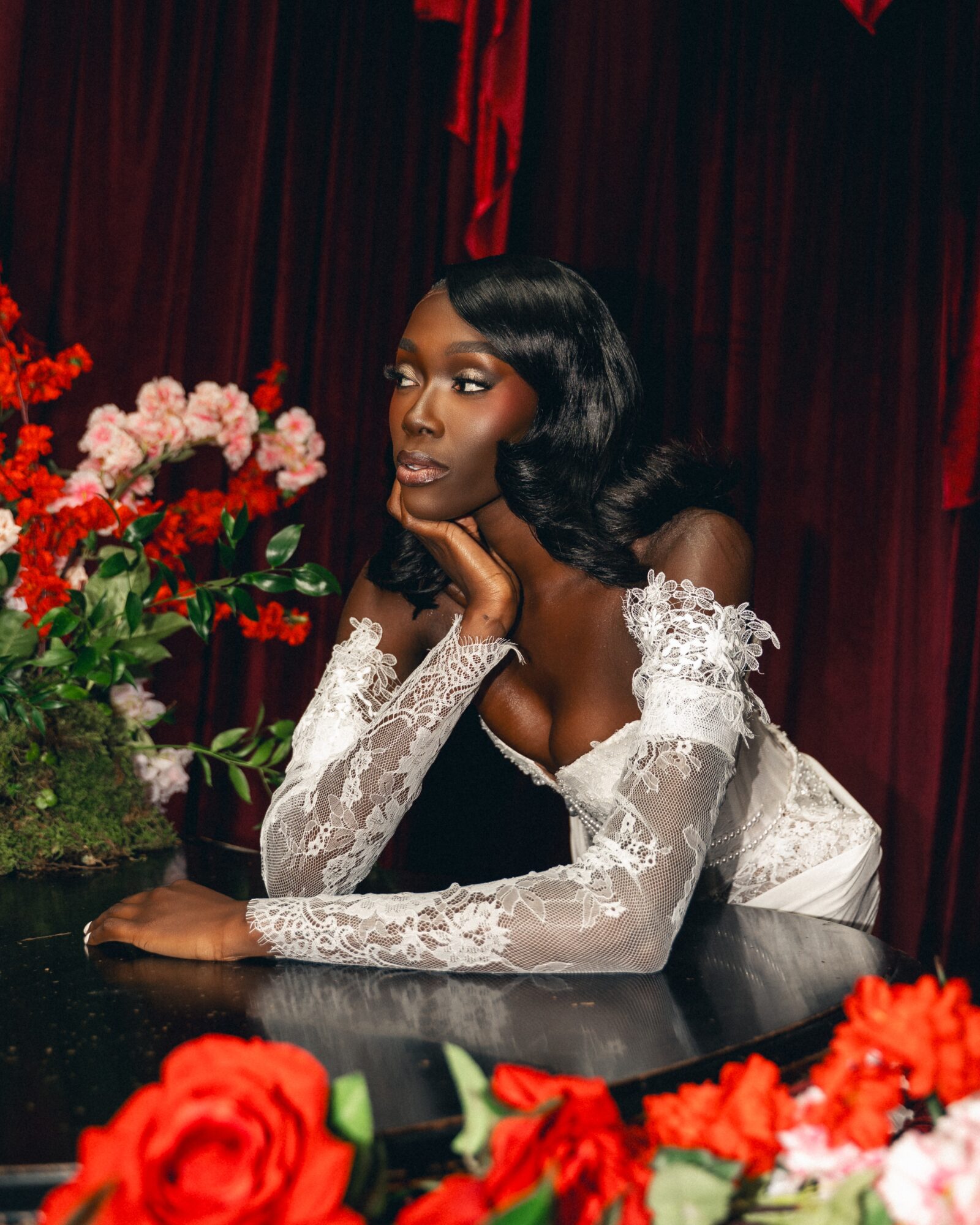 Woman with wavy black hair and lace dress resting her chin on her hand, surrounded by red and pink flowers, dark background.