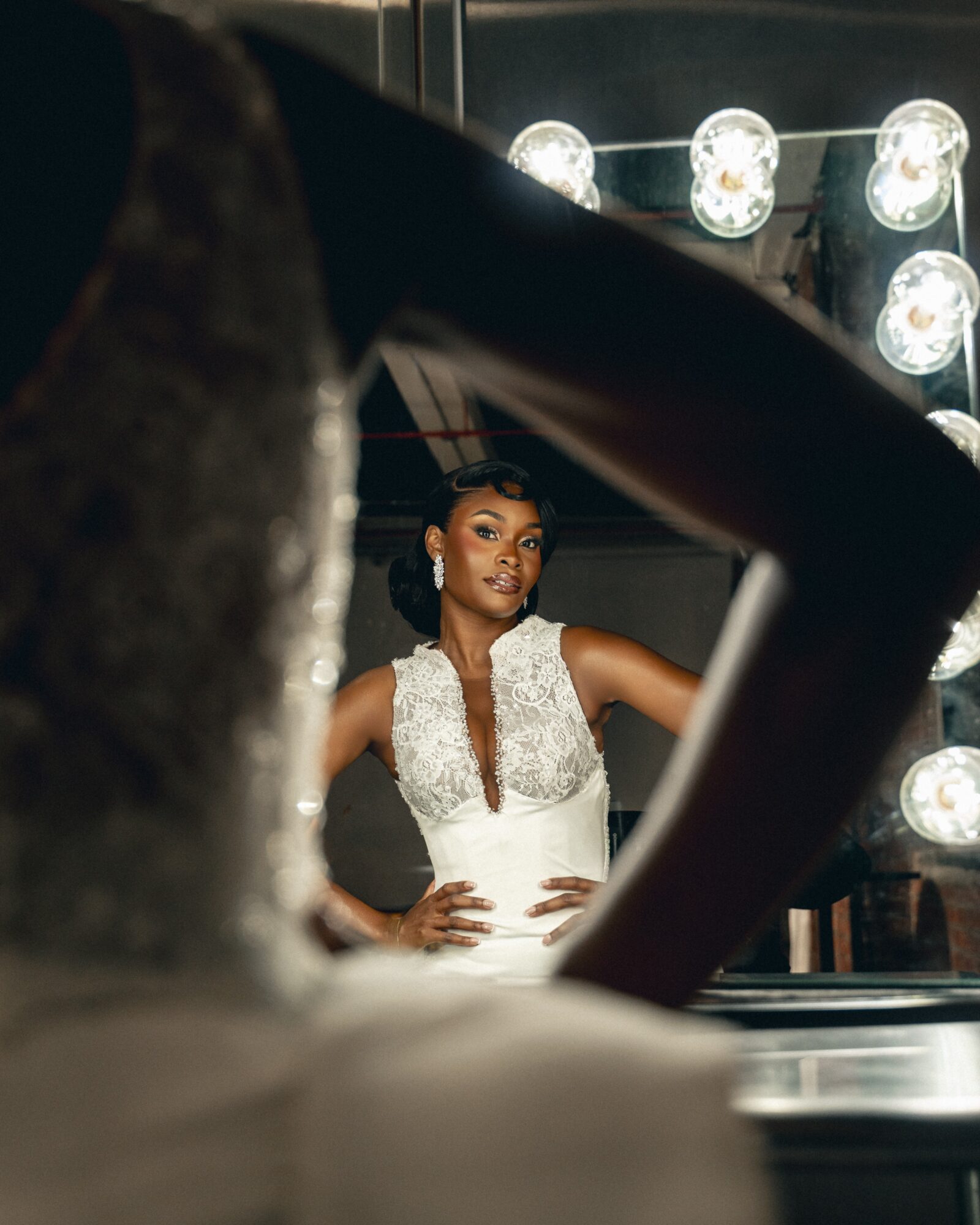 A woman in a white dress with silver embellishments poses with hands on hips, seen through a frame of another person's arm.