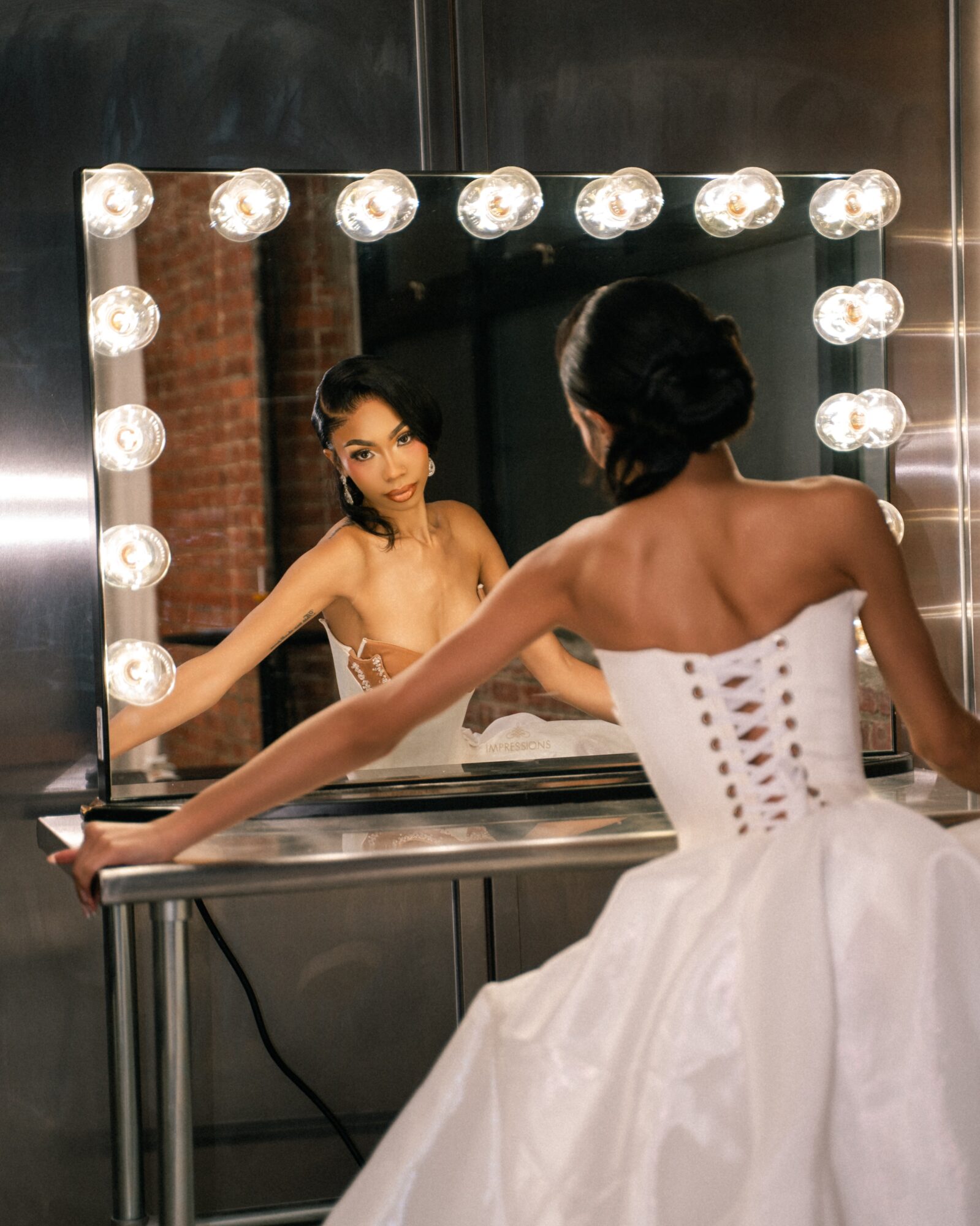 Woman in a white dress looking at her reflection in a mirror with light bulbs, in a dressing room.
