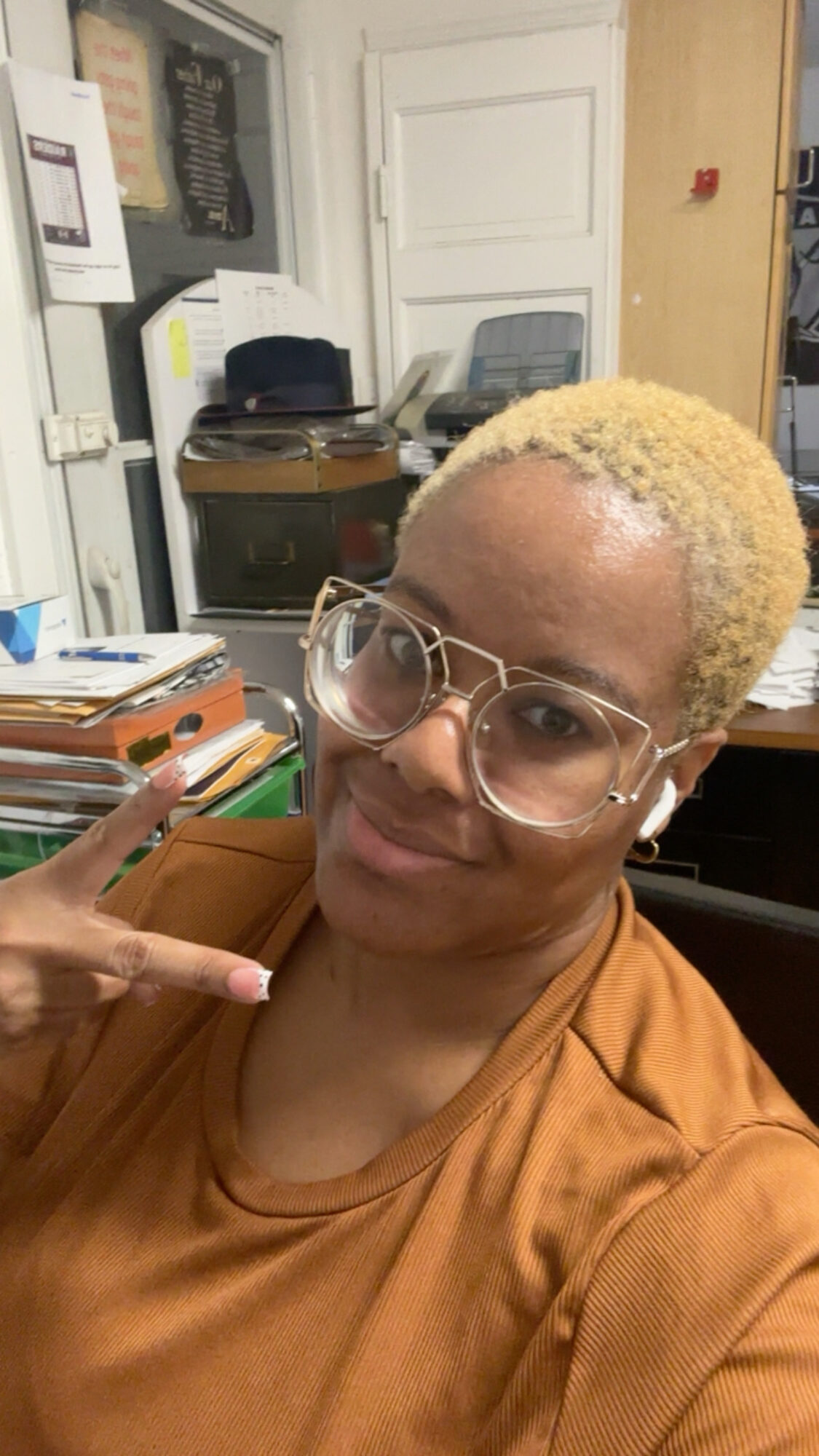 Person with short blonde hair, glasses, and a brown shirt making a peace sign gesture indoors.