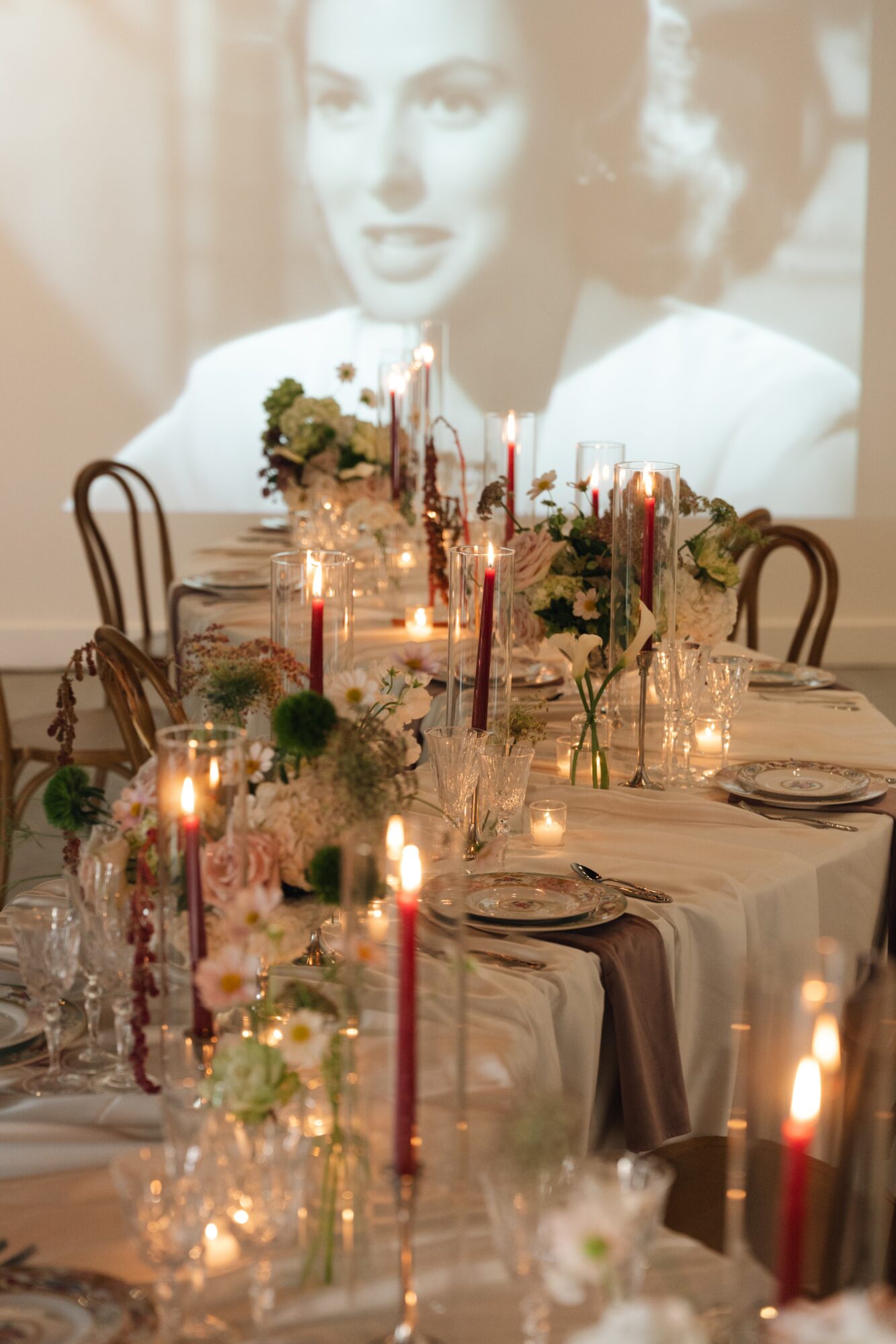 Decorated dining table with candles, flowers, and glassware, with a blurred black-and-white portrait of a woman projected on the wall behind.