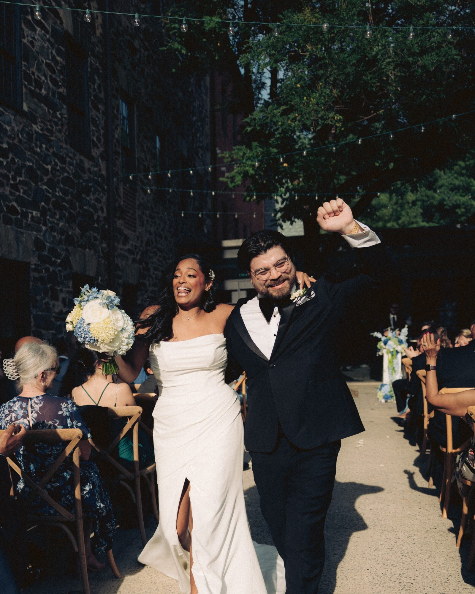 A smiling couple in formal attire walking outdoors, with guests seated on either side, trees, and a stone building in the background.