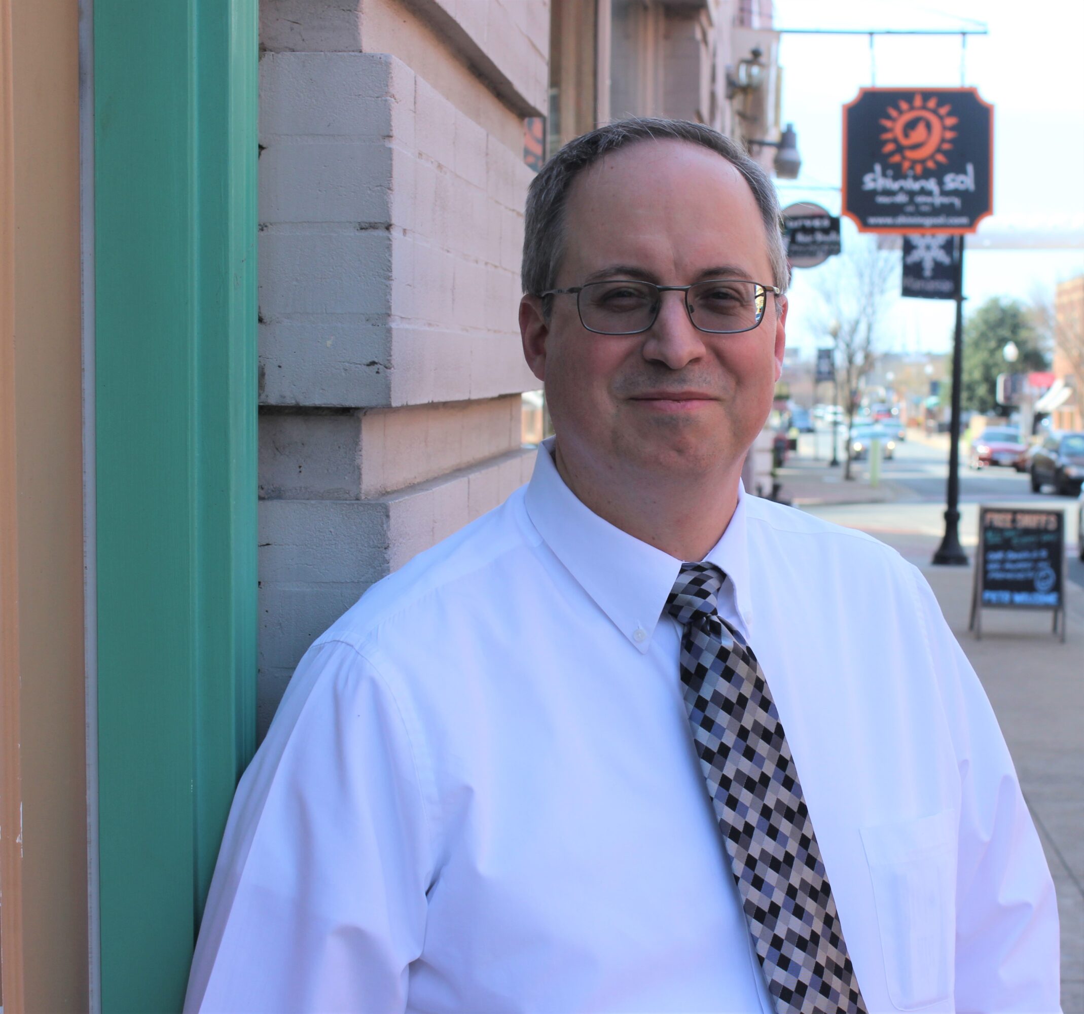 Man in white shirt and patterned tie standing outside near a brick wall, smiling, with street and sign in background.